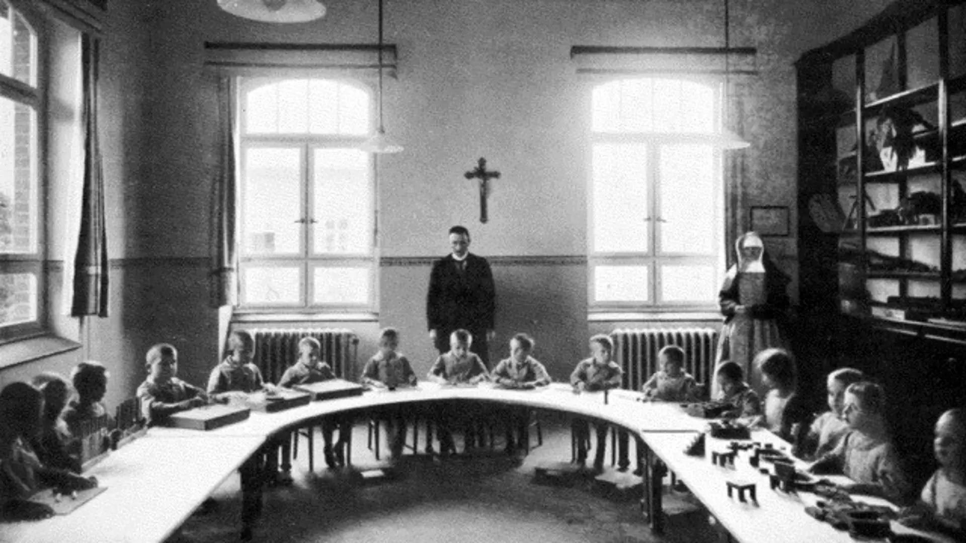  Ein altes, monochromes Foto von einem der Schulräume der Blindenanstalt in den 1920er Jahren. Viele Kinder im Vorschulater sitzen nebeneinander an ihren Plätzen an einem U-förmigen langen Tisch. An der Wand rechts gibt es große Regale mit Büchern, gerade aus und links sind große Fenster zu sehen. Hinter den Schüler*innen stehen ein Priester und zwei Nonnen. Höchstwahrscheinlich sind diese der Klassenlehrer und seine Hilfskräfte. 