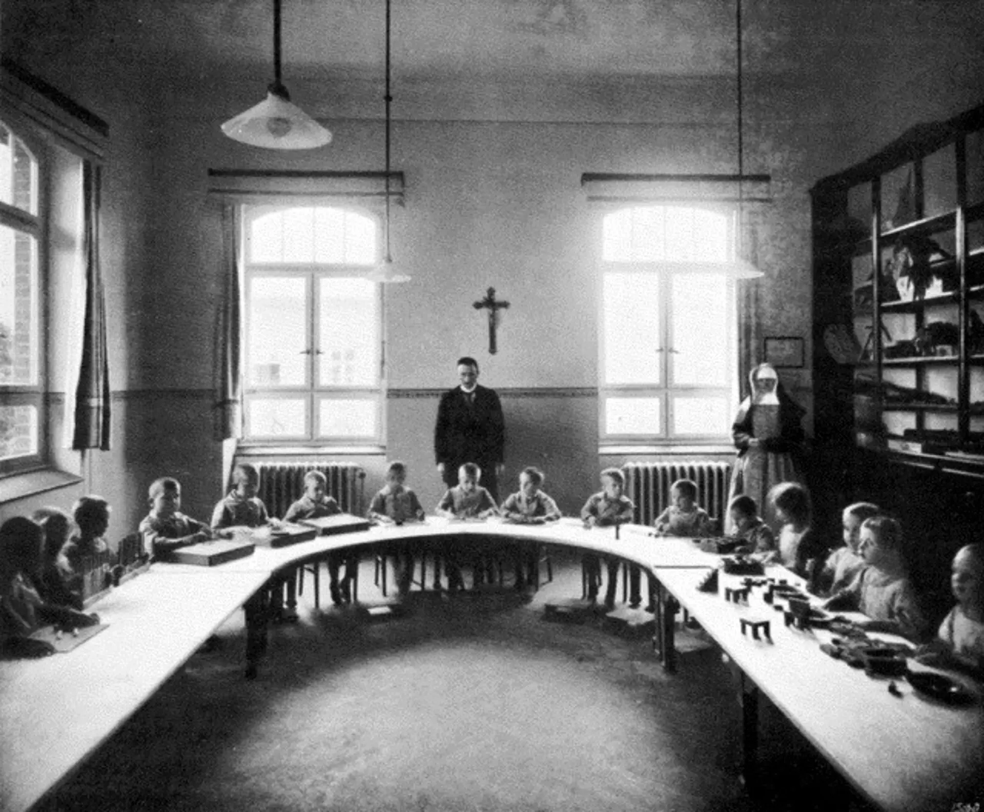 Ein altes, monochromes Foto von einem der Schulräume der Blindenanstalt in den 1920er Jahren. Viele Kinder im Vorschulater sitzen nebeneinander an ihren Plätzen an einem U-förmigen langen Tisch. An der Wand rechts gibt es große Regale mit Büchern, gerade aus und links sind große Fenster zu sehen. Hinter den Schüler*innen stehen ein Priester und zwei Nonnen. Höchstwahrscheinlich sind diese der Klassenlehrer und seine Hilfskräfte. 