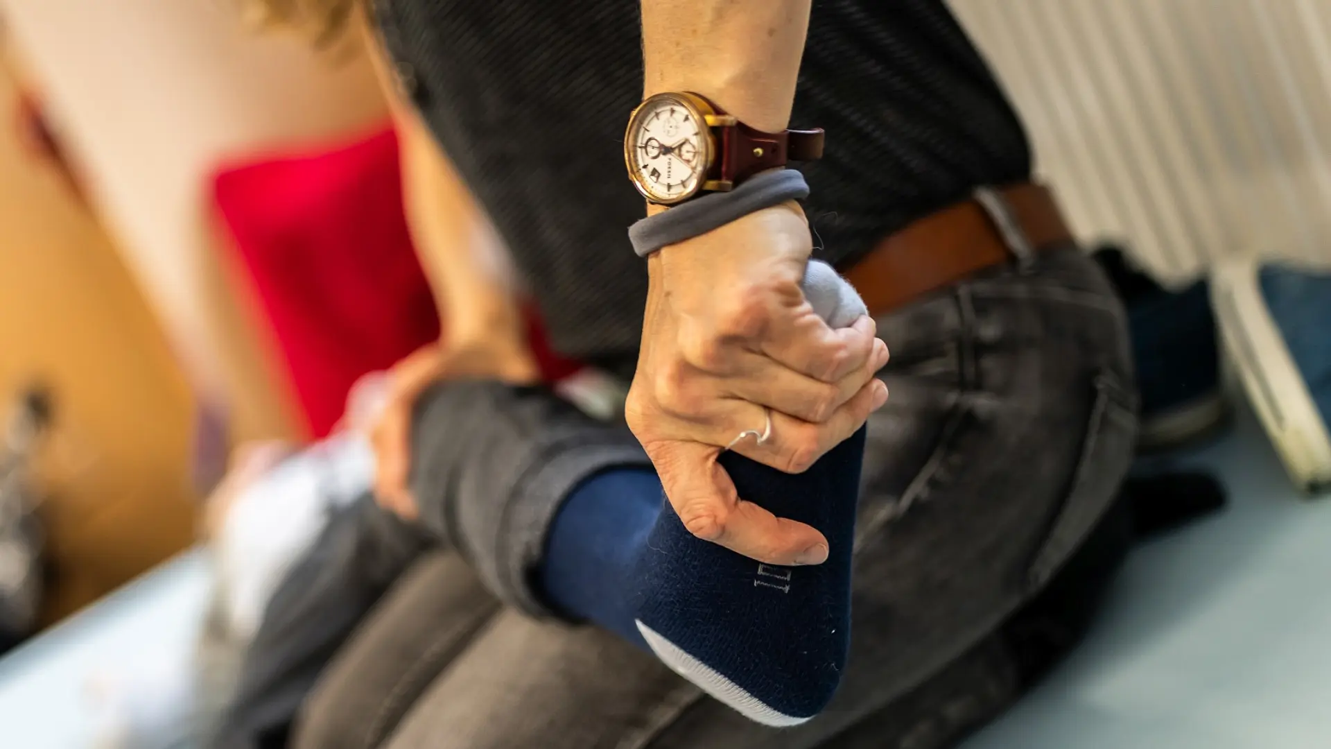 Eine Therapiefachkraft kniet auf einer Matte auf dem Boden, vor ihr liegt ein Kind. Mit der einen Hand hält sie leicht das Knie des Kindes, mit der anderen drückt sie an seinem Fuß.