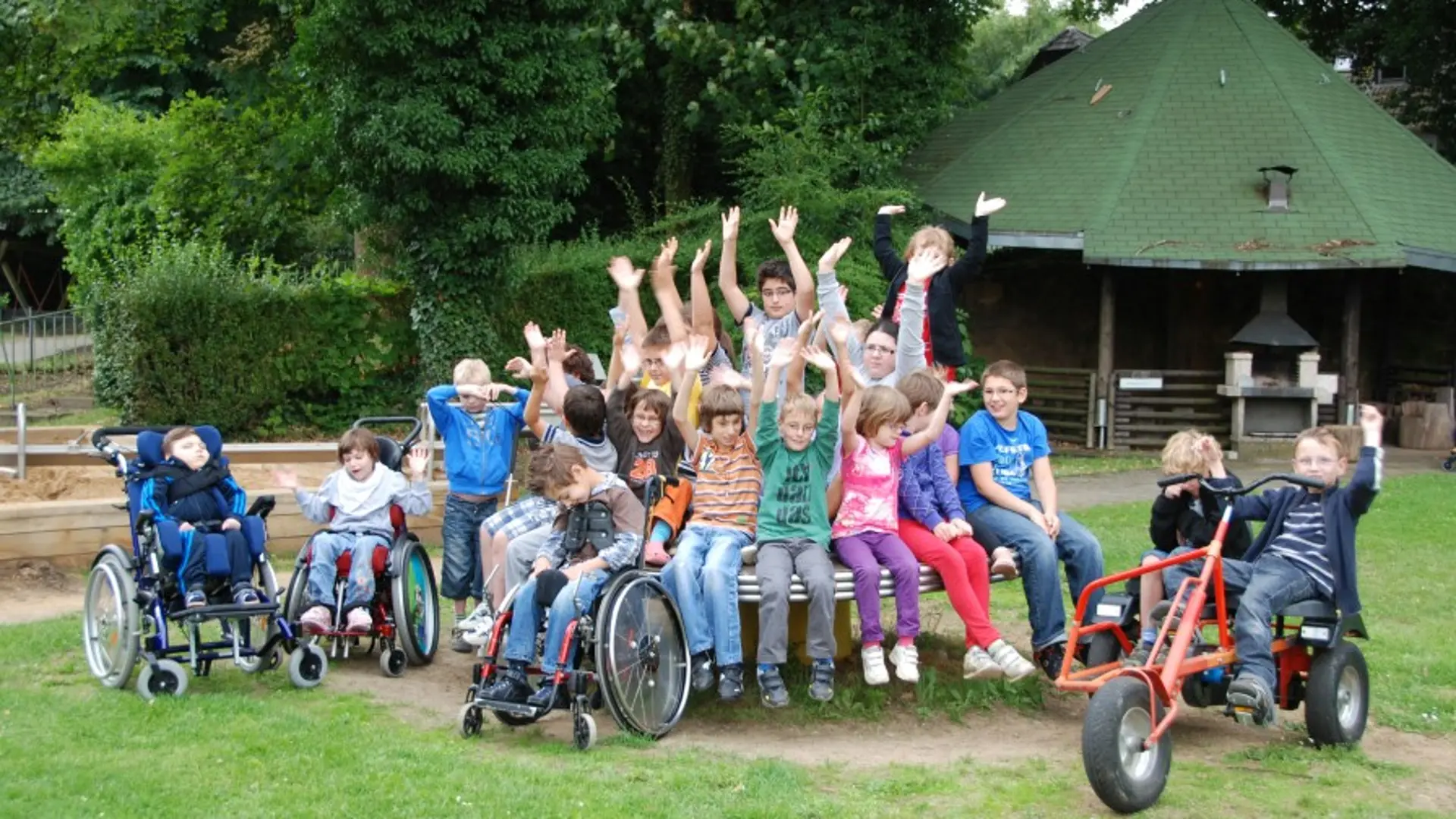 Eine Gruppe mit etwa 20 Kindern im Grundschulalter posiert für ein gemeinsames Foto an einem Spielplatz draußen im Freien. Die Mehrheit der Kinder sitzt dicht nebeneinander auf einem runden Klettertisch aus Metall. Drei Kinder mit komplexer Beeinträchtigung sind im Rollstuhl und stehen neben dem Tisch links. Zwei Schüler in Tretbuggies sind auf der anderen Seite des Tisches. Alle Kinder schauen in die Kamera und manche heben die Arme hoch in eine Art fröhliche Begrüßung.