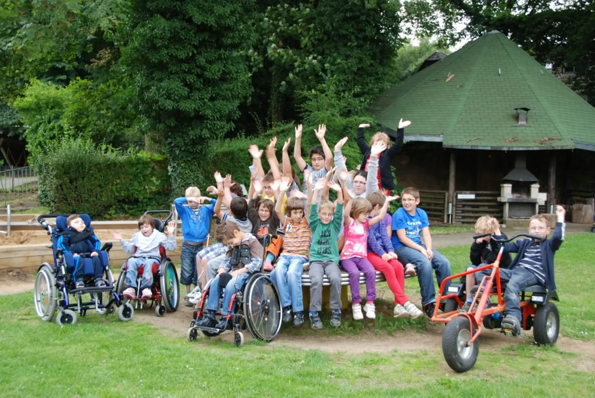 Eine Gruppe mit etwa 20 Kindern im Grundschulalter posiert für ein gemeinsames Foto an einem Spielplatz draußen im Freien. Die Mehrheit der Kinder sitzt dicht nebeneinander auf einem runden Klettertisch aus Metall. Drei Kinder mit komplexer Beeinträchtigung sind im Rollstuhl und stehen neben dem Tisch links. Zwei Schüler in Tretbuggies sind auf der anderen Seite des Tisches. Alle Kinder schauen in die Kamera und manche heben die Arme hoch in eine Art fröhliche Begrüßung.