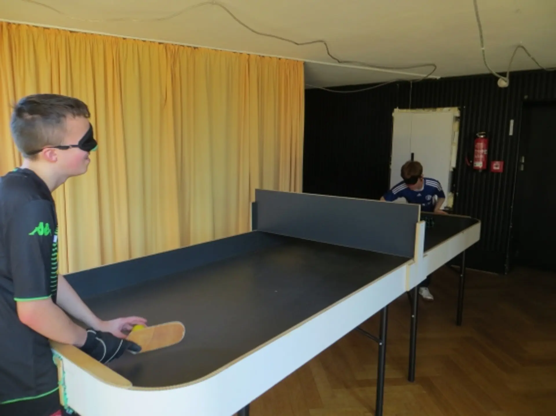 Zwei Schüler spielen Tischball in einem Raum. Der Raum hat keine Fenster auf der Seite, wo der Tisch steht, sondern ein schwerfallender Vorhang. Hinter dem einen Jungen ist auch der Ausgang.