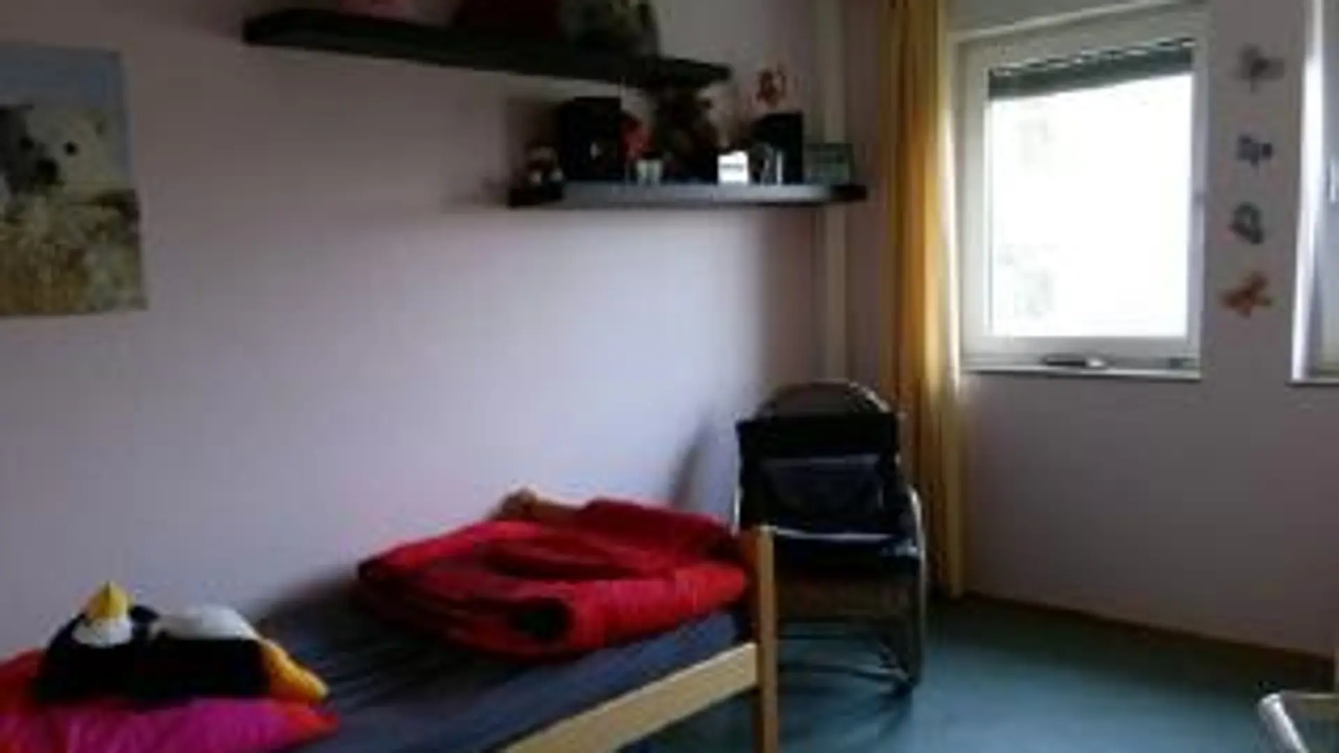 Ein Zimmer im Internat, das farbenfroh eingerichtet ist. Man sieht ein Bett mit frisch bezogenen Kissen und Decke, ein Plüschpinguin liegt auf dem Kissen. An der Wand über dem Bett ist die Ecke eines Posters mit einem Polarbär zu sehen. Neben dem Bett ist noch ein Stuhl. Man sieht noch zwei Fenster mit offenen Gardinen. An der Wand ist noch ein Eckregal befestigt, auf dem persönliche Gegenstände liegen.