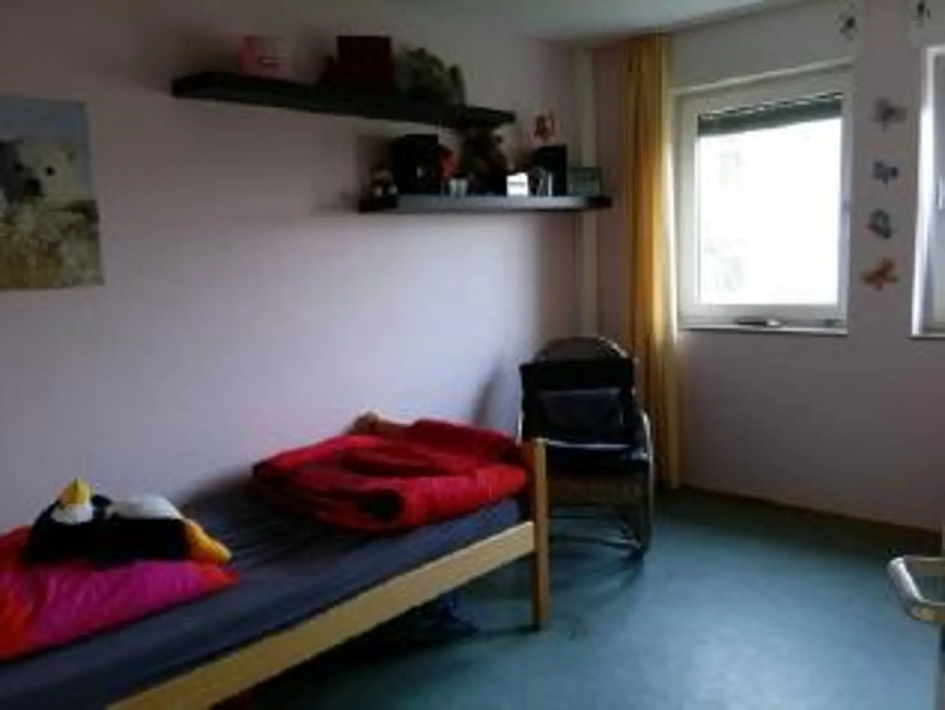 Ein Zimmer im Internat, das farbenfroh eingerichtet ist. Man sieht ein Bett mit frisch bezogenen Kissen und Decke, ein Plüschpinguin liegt auf dem Kissen. An der Wand über dem Bett ist die Ecke eines Posters mit einem Polarbär zu sehen. Neben dem Bett ist noch ein Stuhl. Man sieht noch zwei Fenster mit offenen Gardinen. An der Wand ist noch ein Eckregal befestigt, auf dem persönliche Gegenstände liegen.