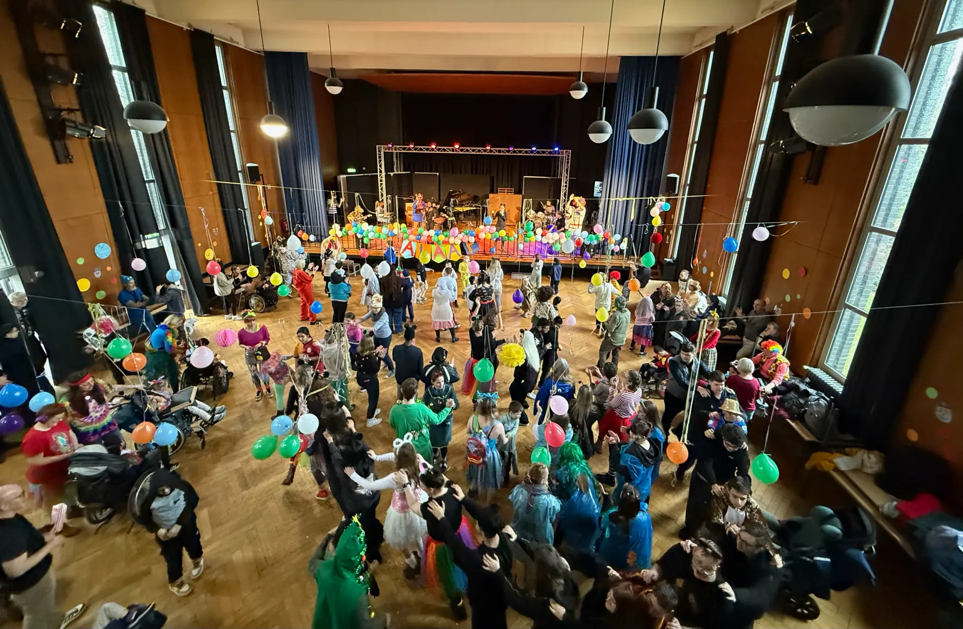 Auf dem Foto ist die mit bunten Luftballons und Girlanden geschmückte Aula zu sehen. Auf der Bühne spielt die Band ein Lied. Vor der Bühne tanzen viele Schüler*innen, Lehrkräfte und weitere Mitarbeitende der Schule.