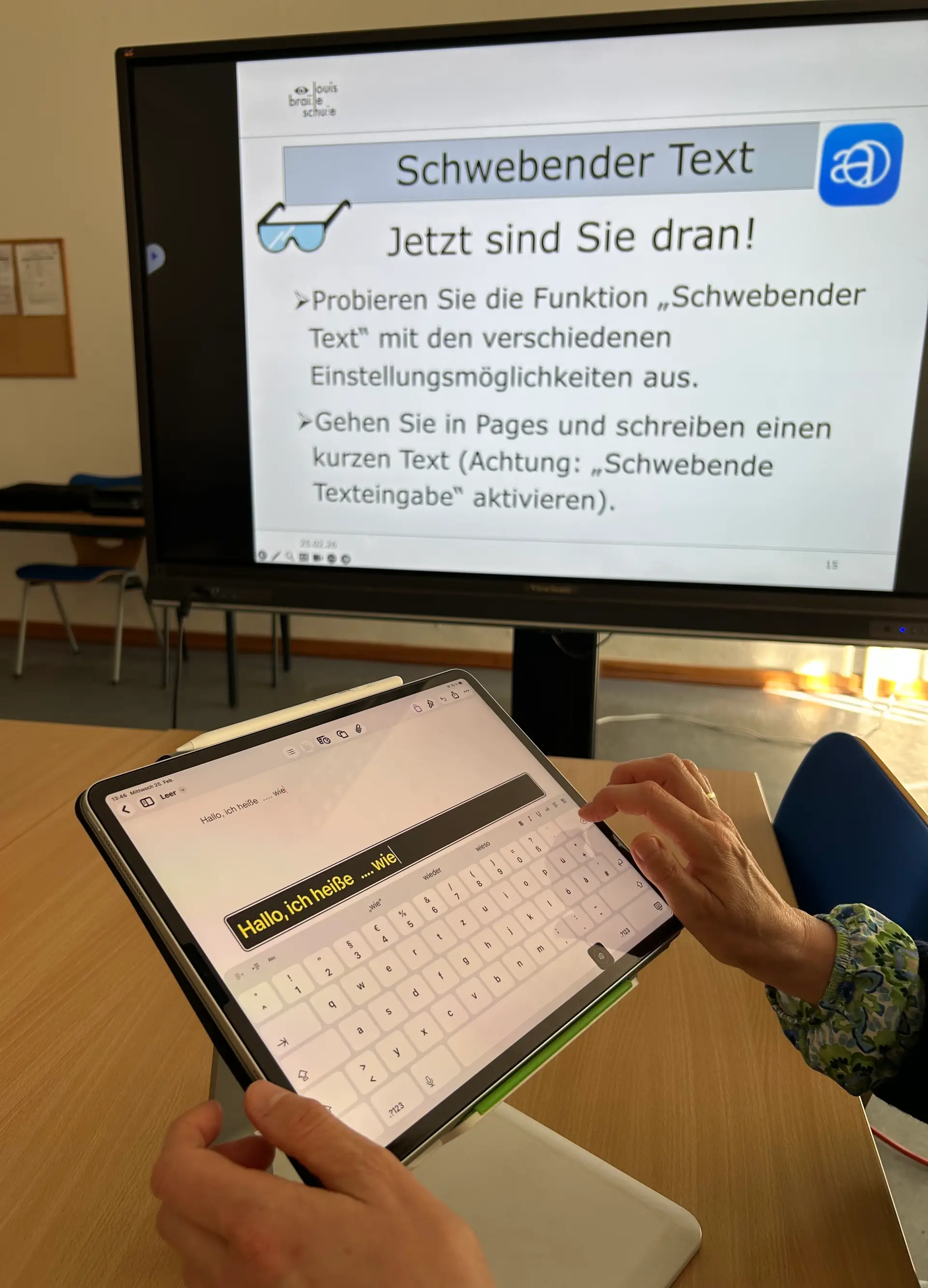 Auf dem Foto ist ein iPad zu sehen, auf welchem die App „Pages“ geöffnet ist. Es sind Hände zu sehen, die auf der iPad-internen Tastatur einen Text in „Pages“ schreiben. Über der Tastatur ist ein Feld zu sehen, welches das Geschriebene vergrößert und in anderen Farben darstellt. Im Hintergrund ist eine digitale Tafel mit einer PowerPoint-Folie mit der Überschrift „Schwebender Text“.