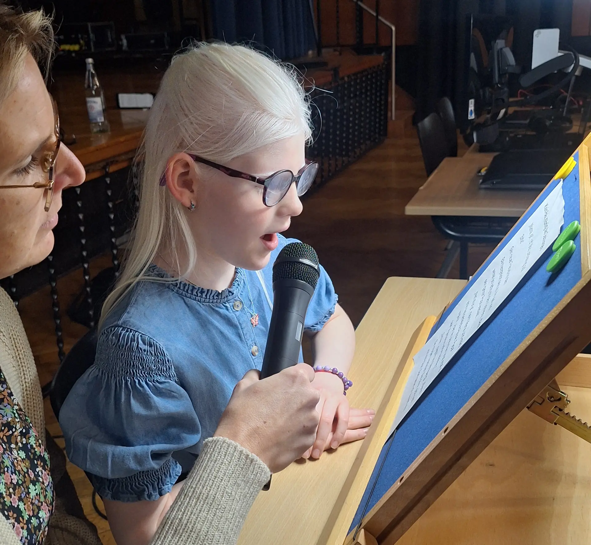 Eine Schülerin mit Brille sitzt an einem Tisch, vor sich ein Lesepult mit ihrem ausgedruckten Text. Eine Leuchte gibt Licht. Neben ihr eine Lehrerin, die ein Mikrofon für die Schülerin hält.