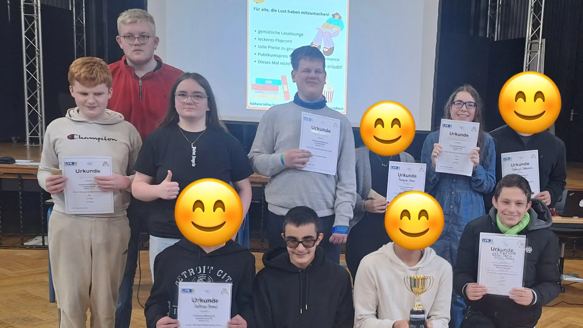 Auf dem Foto sind insgesamt sieben Schüler*innen, zu einem Gruppenfoto aufgestellt, zu sehen. Sie halten Urkunden sowie einen Pokal. Vier Gesichter sind mit Smileys abgedeckt. Im Hintergrund ist die Leinwand mit dem Plakat des Lesewettbewerbs zu sehen.