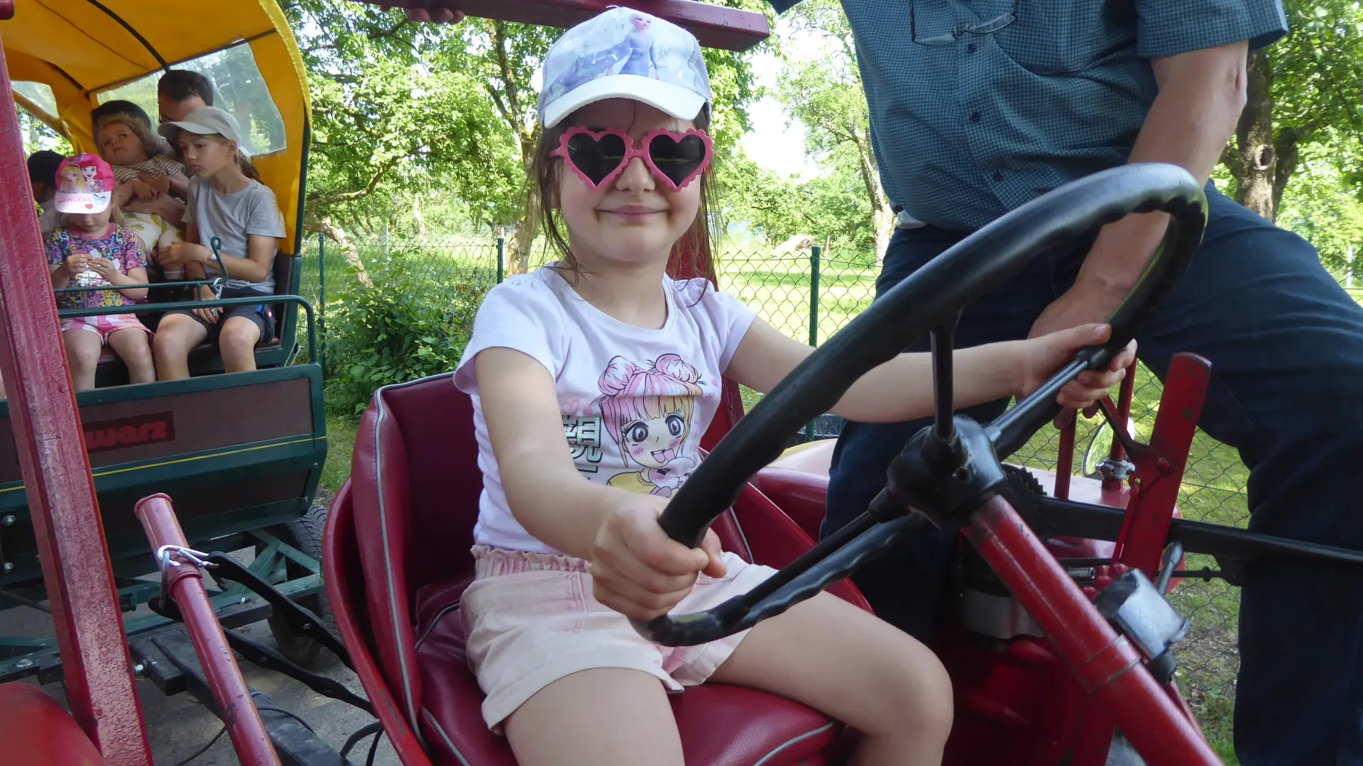 Ein Mädchen sitzt auf einem Traktor. Im Hintergrund sieht man die Familien im Planwagen.