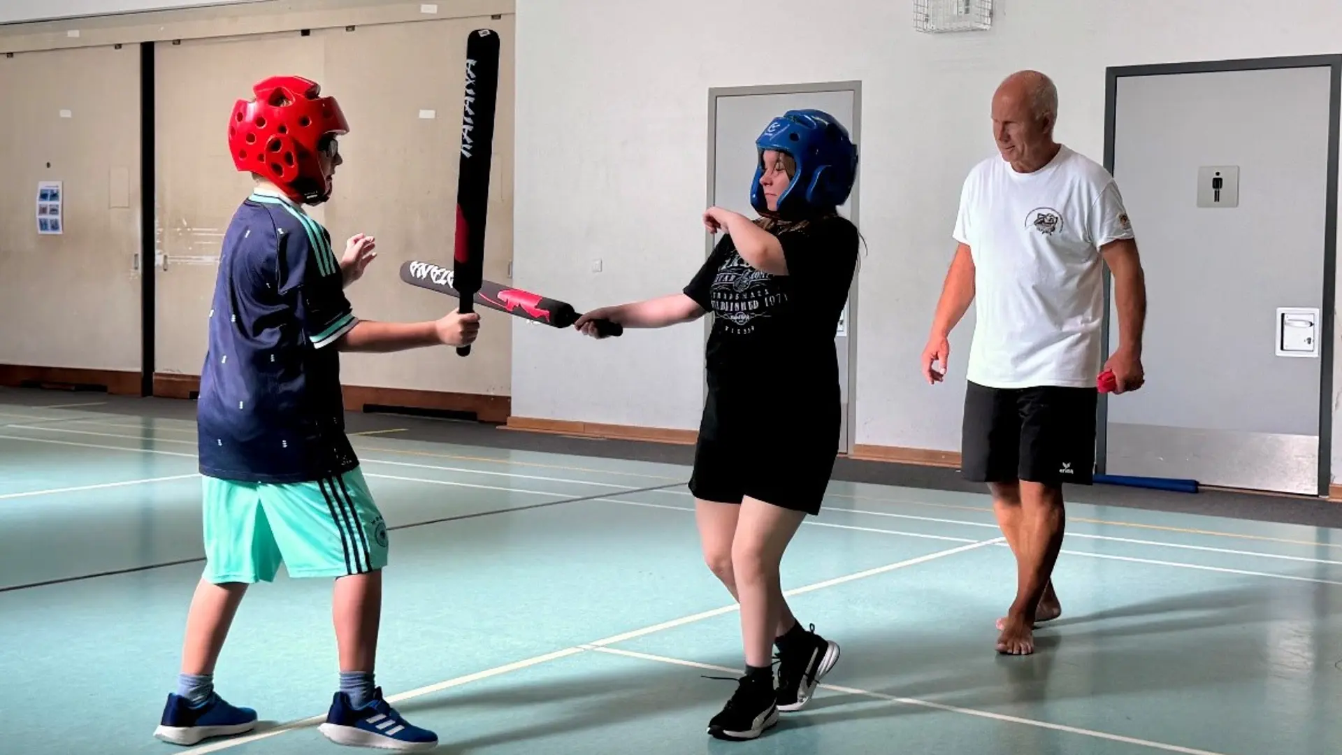 Zwei Mädchen in der Turnhalle mit Helmen und Schaumstoffschlägern. Im Hintergrund steht ein Mann und beobachtet die beiden. 
