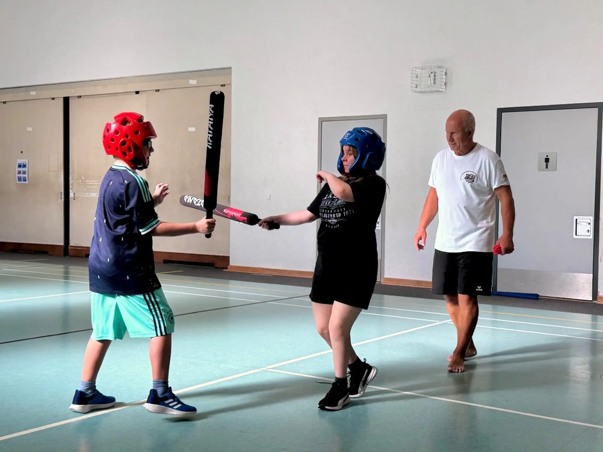 Zwei Mädchen in der Turnhalle mit Helmen und Schaumstoffschlägern. Im Hintergrund steht ein Mann und beobachtet die beiden.