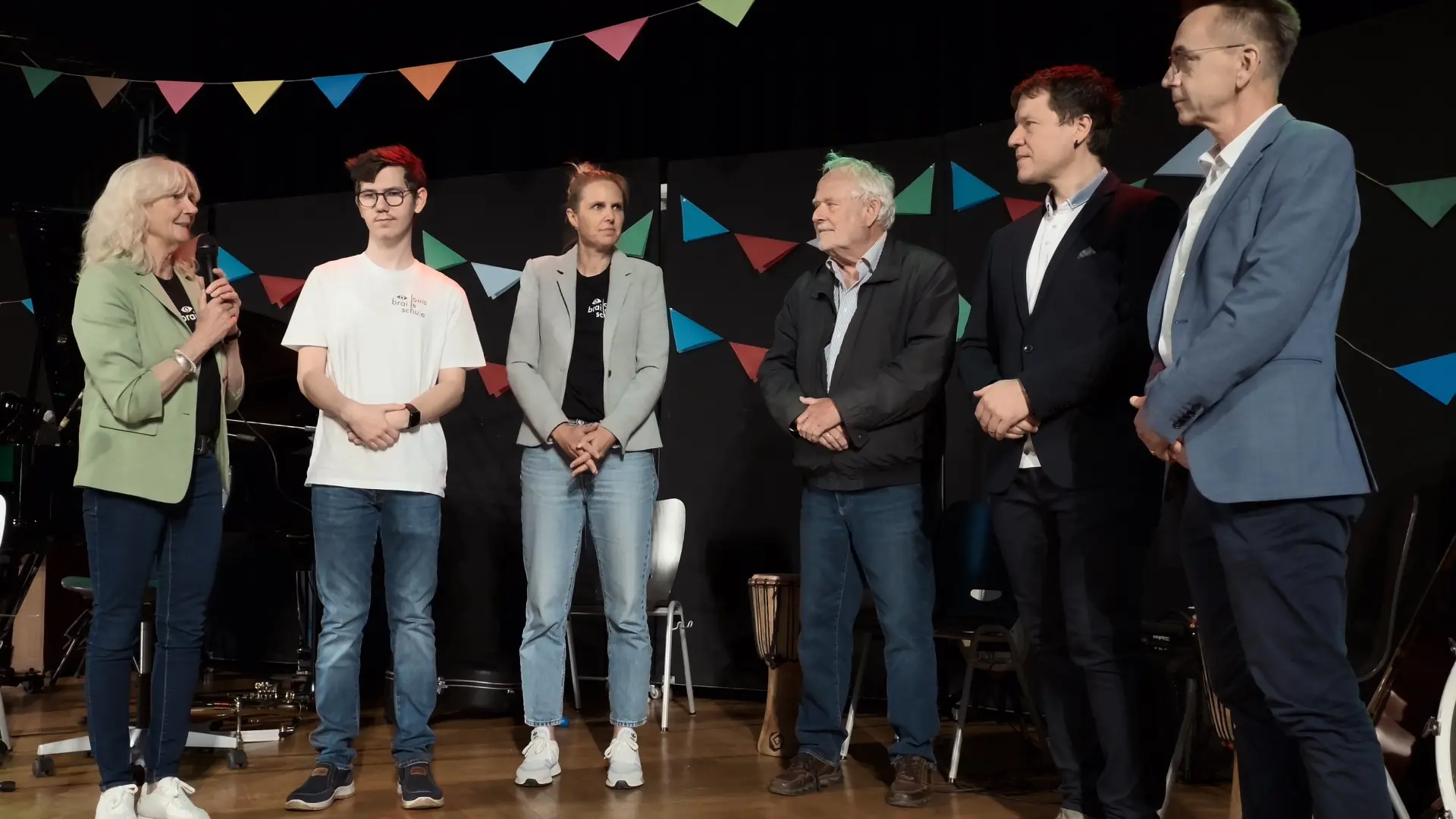 Auf der Bühne stehen sechs Personen (u.a. Schulleiterin Frau Grün-Klingebiel, Schülersprecher David Jelic und der Vorsitzende des Fördervereins, Herr Reith). Sie würdigten die wertvolle Unterstützung zahlreicher Unterstützer*innen der Schule. Frau Grün-Klingebiel hält das Mikrofon.