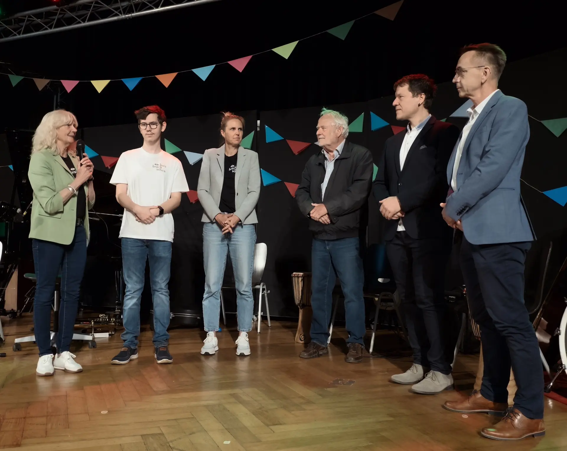 Auf der Bühne stehen sechs Personen (u.a. Schulleiterin Frau Grün-Klingebiel, Schülersprecher David Jelic und der Vorsitzende des Fördervereins, Herr Reith). Sie würdigten die wertvolle Unterstützung zahlreicher Unterstützer*innen der Schule. Frau Grün-Klingebiel hält das Mikrofon.
