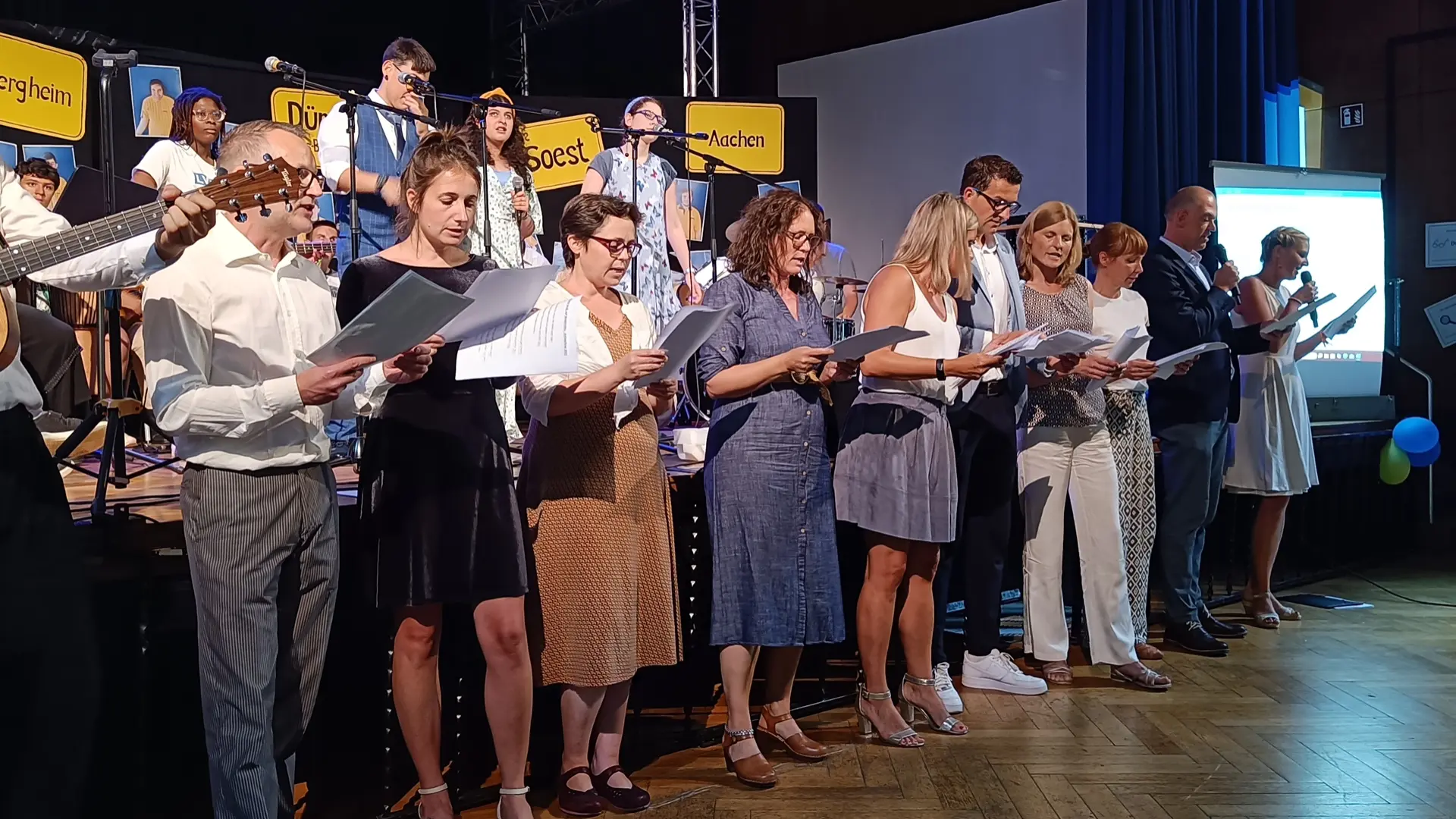 Foto der Abschlussfeier 2025 der LVR-Louis-Braille-Schule. Viele Personen stehen mit Notenblättern in den Händen vor der Bühne und singen. Einige Personen musizieren auf Instrumenten.
