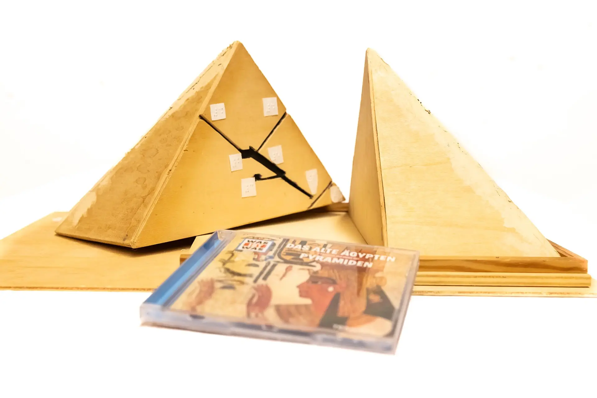 Modell einer ägyptischen Pyramide aus hellbraunem Holz, sodass dadurch die Farbe des Wüstensands imitiert wird. Das Modell lässt sich in der Mitte trennen und auf der einen Hälfte sind die inneren Gänge in Braille-Schrift gekennzeichnet. Zur Pyramide liegt eine Hörbuch-CD von Was ist Was mit dem Titel Das alte Ägypten. Pyramiden.