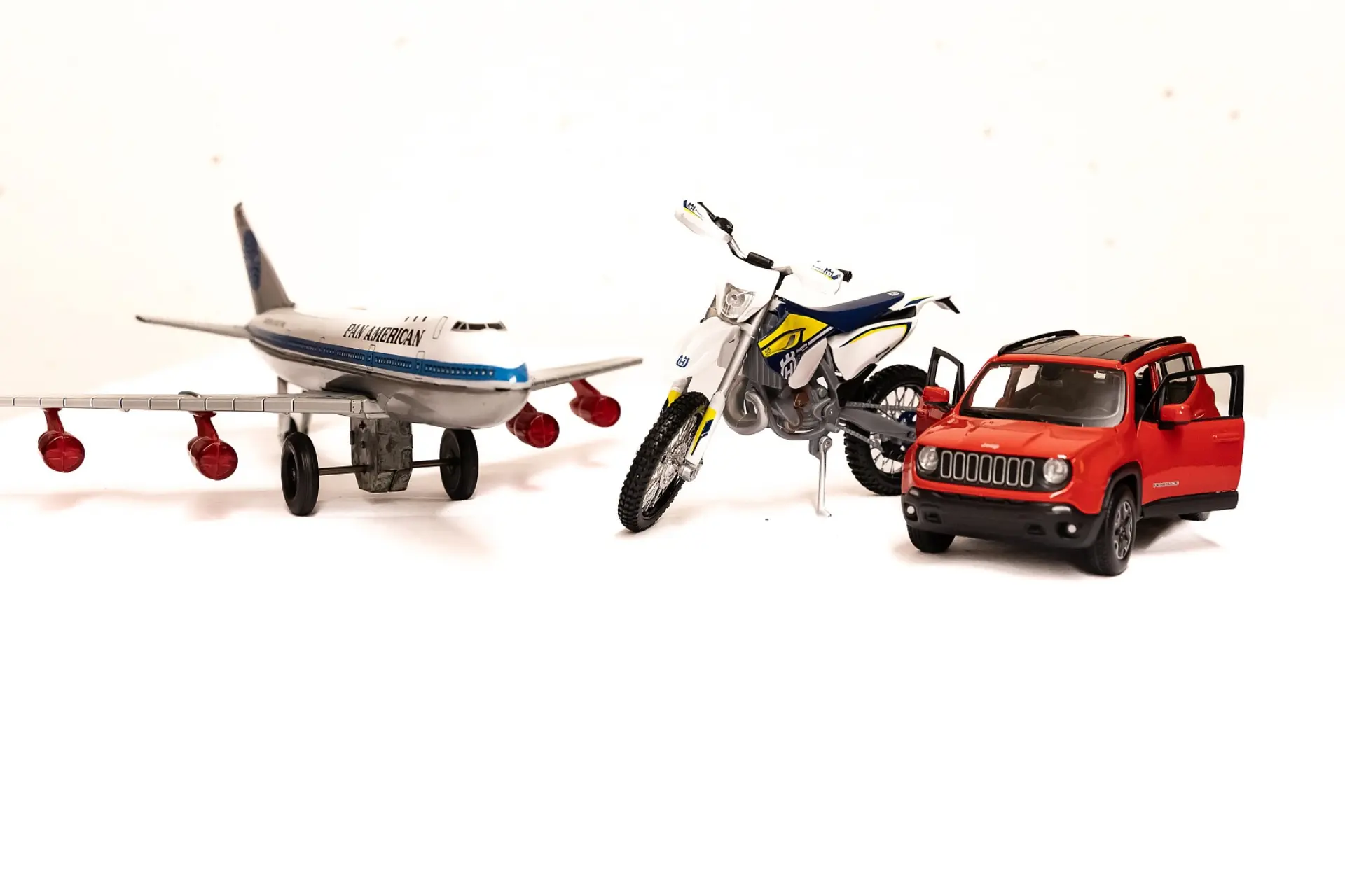 Drei taktile Modellfahrzeuge auf einem weißen Hintergrund arrangiert. Von links nach rechts sind zu sehen: ein großes Passagierflugzeug mit der Aufschrift 'PAN AMERCAN' auf dem Rumpf, eine Motocross-Maschine mit detaillierten Rädern und Lenker, und ein Geländewagen mit geöffneten Türen.
