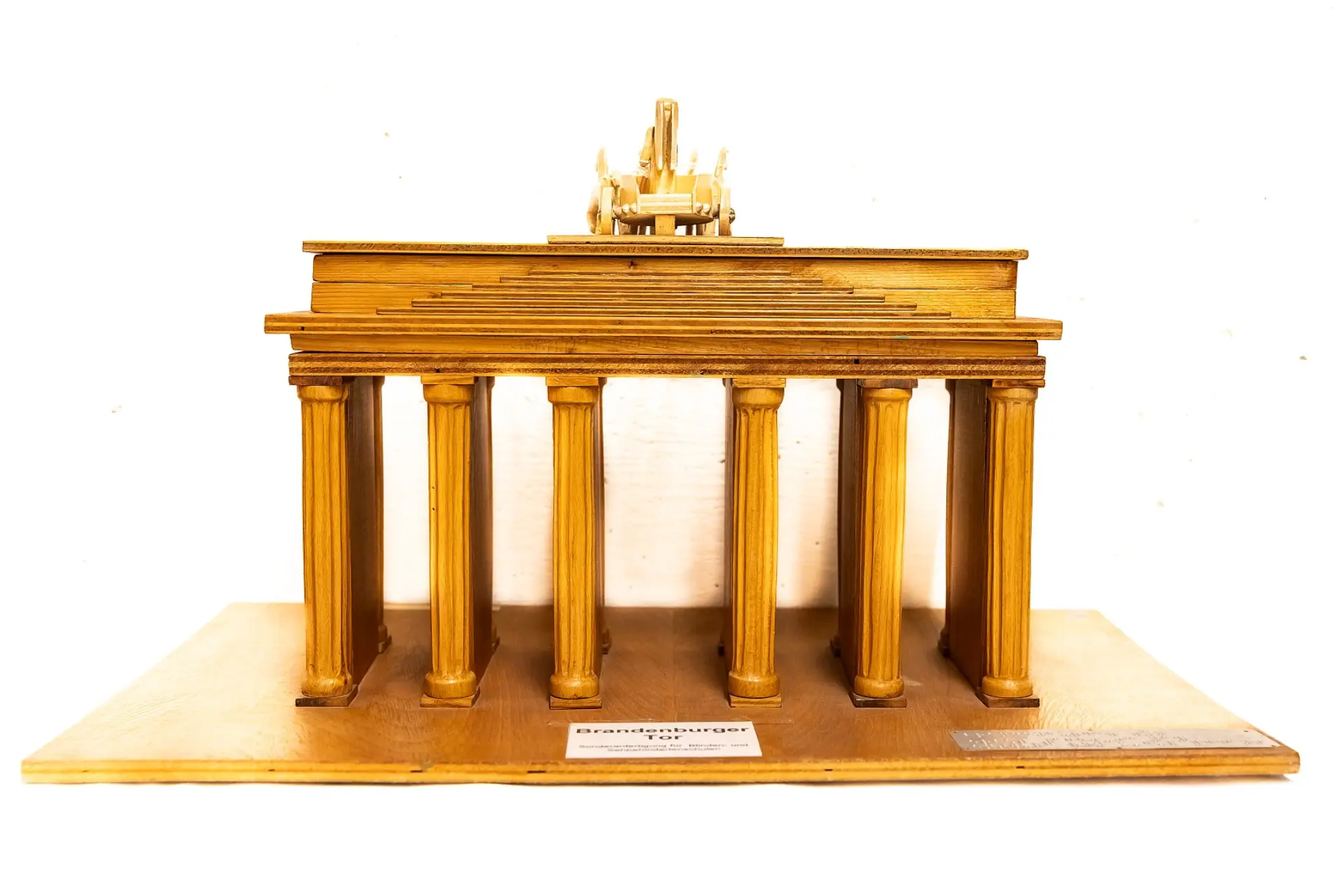 Ein detailliertes, hölzernes Tastmodell des Brandenburger Tors, das auf einer rechteckigen Holzplatte steht. Das Tor zeigt die charakteristischen Säulen und den oberen Fries. Oben auf dem Tor ist die Quadriga, das von Pferden gezogene Streitwagen-Ensemble, erkennbar. Die Grundplatte trägt eine Gravur mit der Aufschrift 'Brandenburger Tor'.