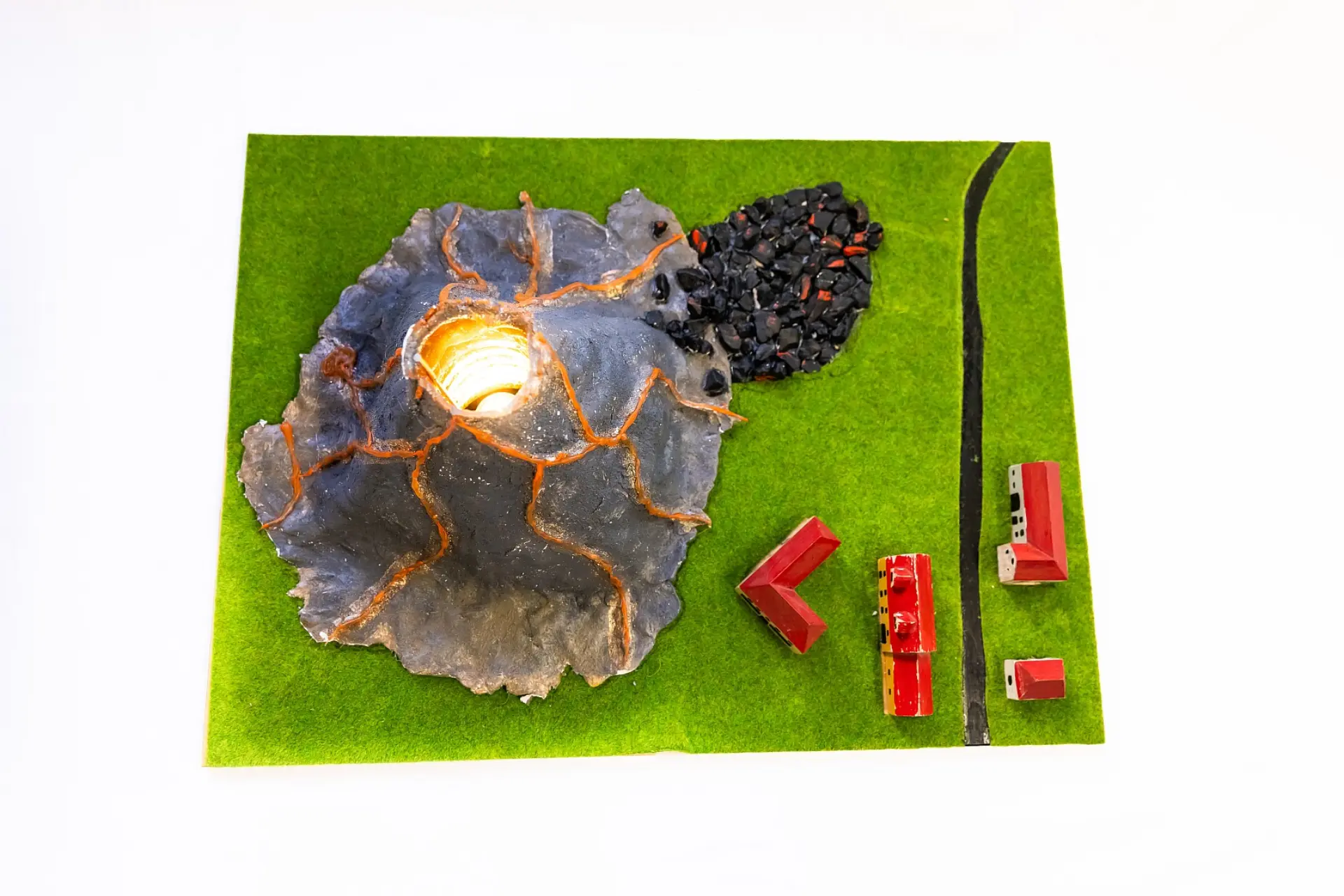Ein taktiles Modell eines Vulkans auf einer Kunstrasen-Grundplatte. Der Vulkan hat orangefarbene Risse, die eine innere Beleuchtung erkennen lassen, die Magma darstellt. Daneben liegt ein Haufen körniger Vulkangesteine. Im unteren Bereich des Modells befinden sich mehrere kleine Häuser und eine dicke Straße, die auf dem helleren Untergrund angedeutet ist.