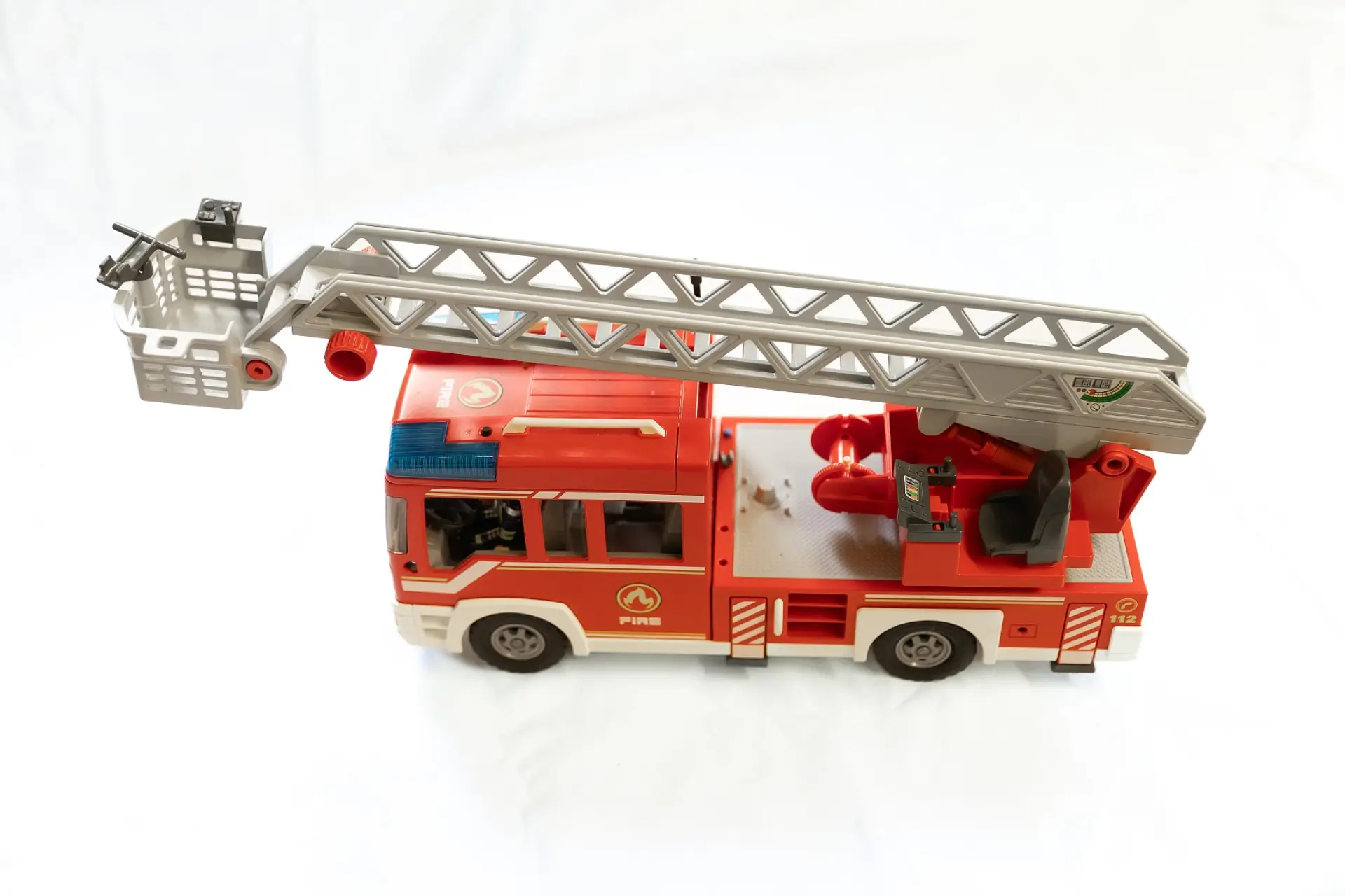 Ein Spielzeug-Feuerwehrauto mit ausfahrbarer Leiter, auf einem weißen Hintergrund platziert. Das Modell hat eine detaillierte Fahrerkabine, Aufschriften wie 'FIRE' und '112' sowie Logos. Die Leiter auf dem Dach ist vollständig ausgefahren und endet in einem Korb mit einem Wasserwerfer. Das Fahrzeug ist stabil gebaut und besitzt funktionale Räder.