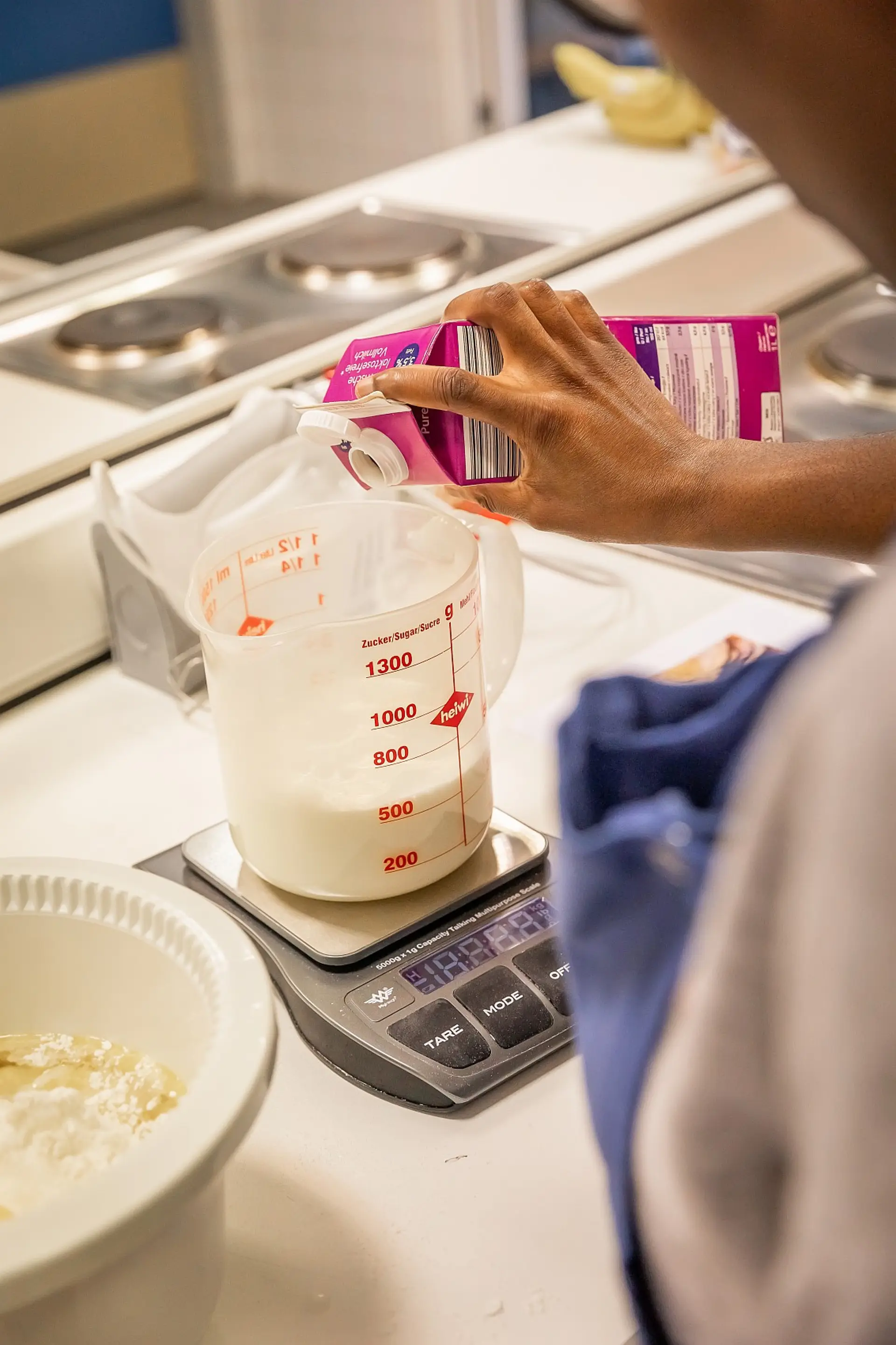 Eine Person bereitet einen Teig in der Schulküche vor und gießt Milch aus dem Tetrapack in einen Messbecher. Der Becher steht auf einer elektrischen sprechenden Waage.
