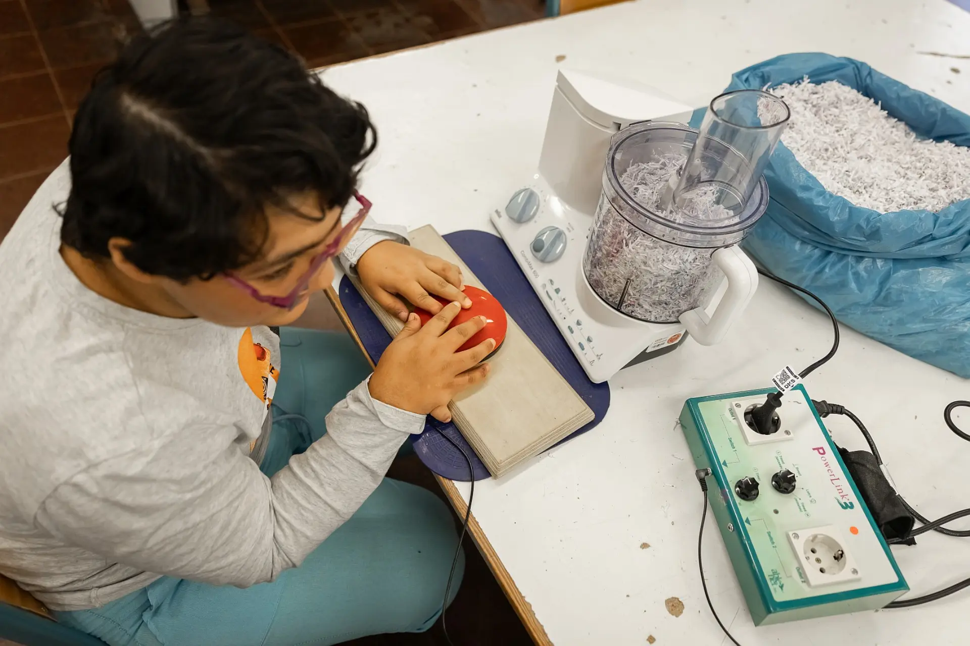 Ein Schüler bedient mithilfe eines handgroßen Knopfes, der mit einem externen Steuergerät verbunden ist, einen elektrischen Zerkleinerer in der Schulküche.