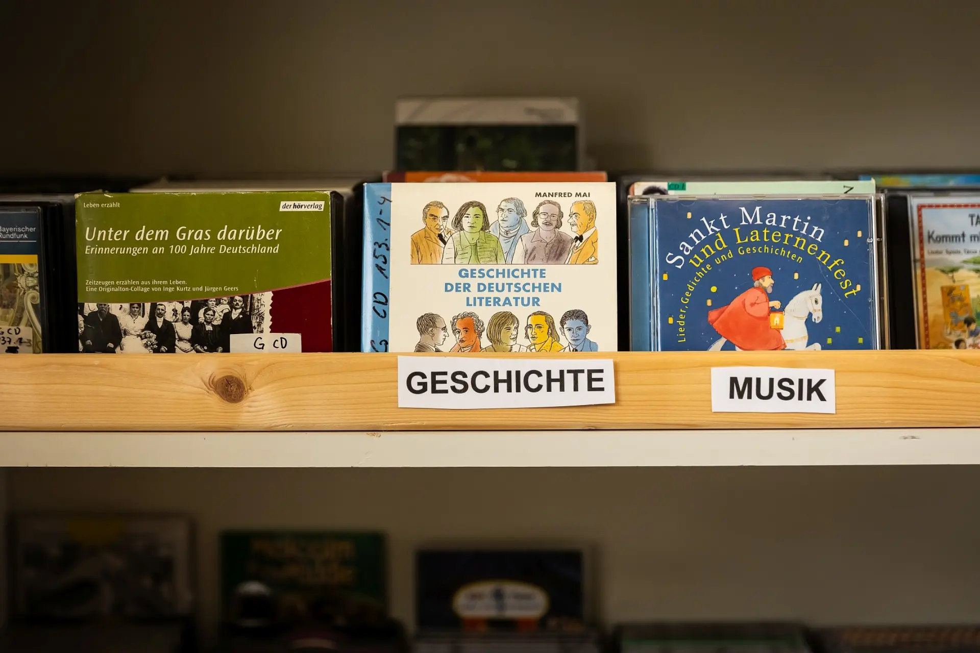Regal mit mehreren CD-Reihen darauf. Die CDs sind mit Hörbüchern für Kinder und Jugendlichen und sie sind nach Genres sortiert. Vor einer CD-Reihe klebt der Zettel "Geschichte", vor der Reihe daneben der Zettel "Musik"