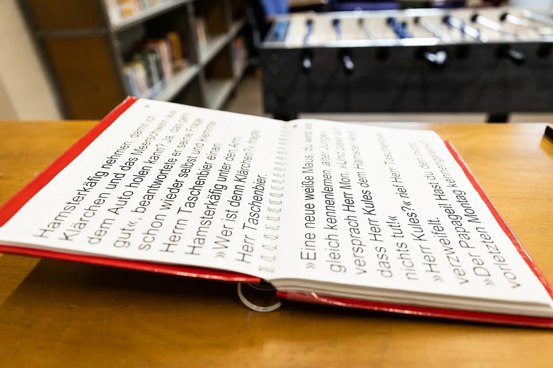 Ein aufgeblättertes Buch mit weißen Seiten und Text mit extra großen Buchstaben in Schwarz liegt auf einem Holztisch. Das Buch wird von der Seite betrachtet, im Hintergrund sieht man noch ein Bücherregal und einen Kickertisch.