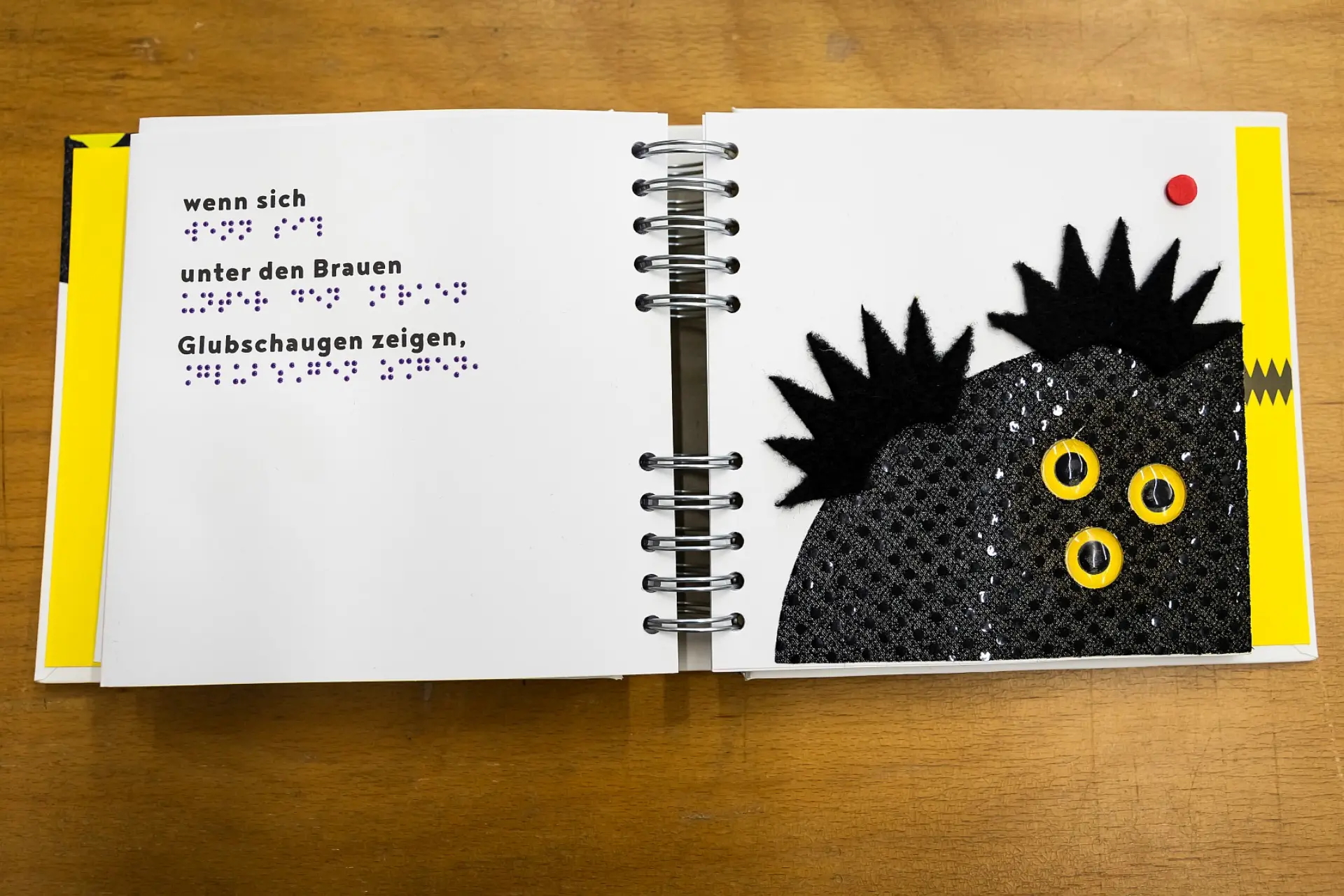Ein aufgeblättertes Buch für Kleinkinder liegt auf einem Tisch. Auf der linken Seite ist der Text Wenn sich unter den Braunen Glubschaugen zeigen sowohl in schwarzen Buchstaben, als auch in Brailleschrift geschrieben. Auf der rechten Seite ist ein Bild von einer Kreatur mit drei Augen, flauschigen Haaren und genopptem Körper, damit die Kinder diese ertasten können. 