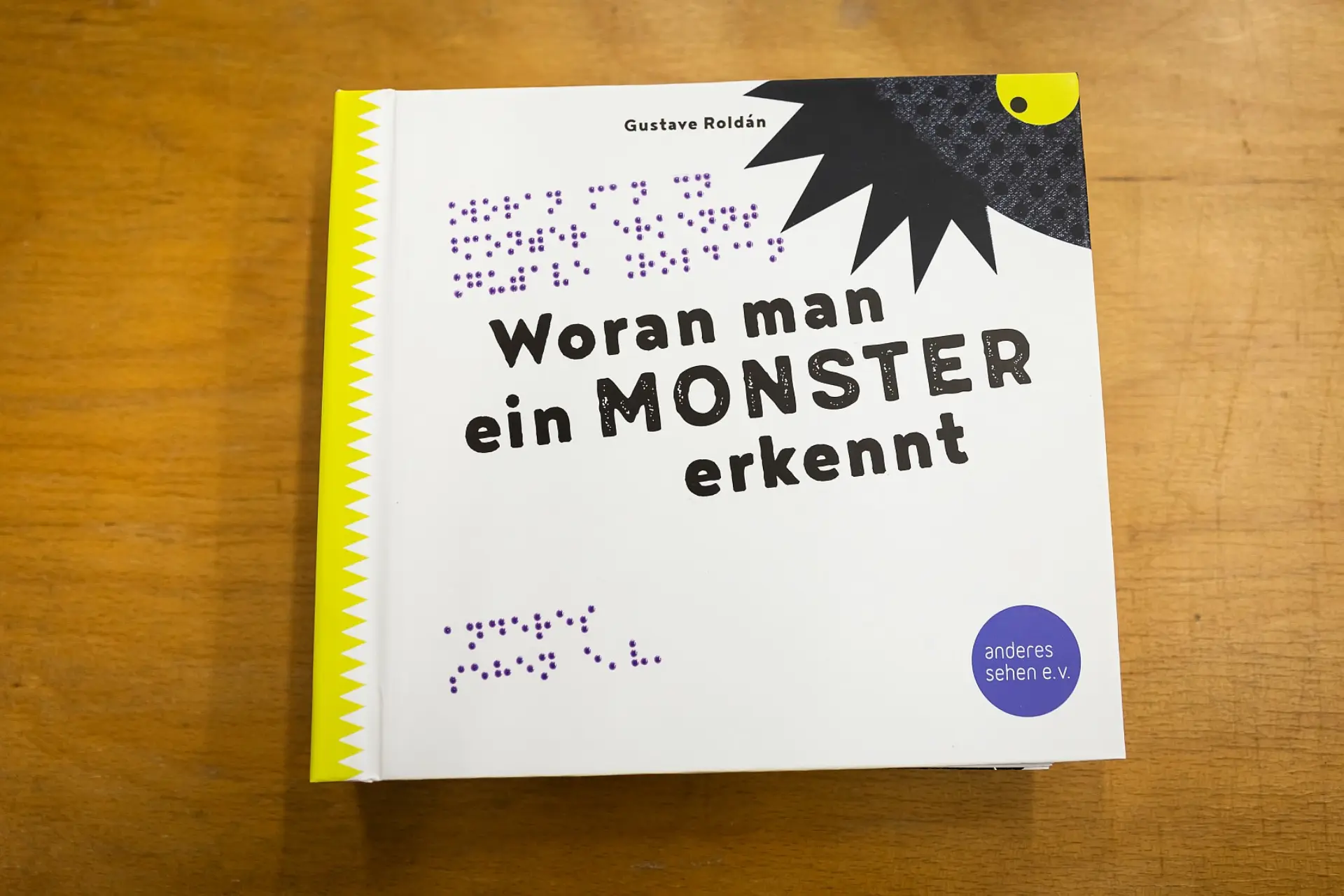 Ein Kinderbuch liegt auf einem Holztisch, nur das Cover ist zu sehen. Das Buch heißt Woran man ein Monster erkennt von Gustave Roldán. Der Verlag heißt anderes sehen ev. Diese Informationen sind auch in Brailleschrift auf dem Cover geschrieben.