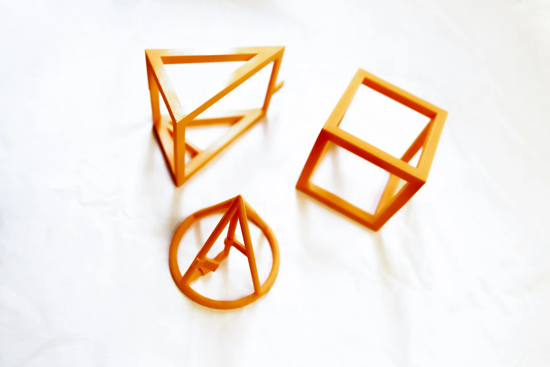 Drei geometrische Körpermodelle als 3D-Figuren (Würfel, Kegel und Dreiecksprisma) mit offener Gitterstruktur, sodass Menschen mit Blindheit oder Sehbeeinträchtigung damit geometrische Aufgaben lösen können.