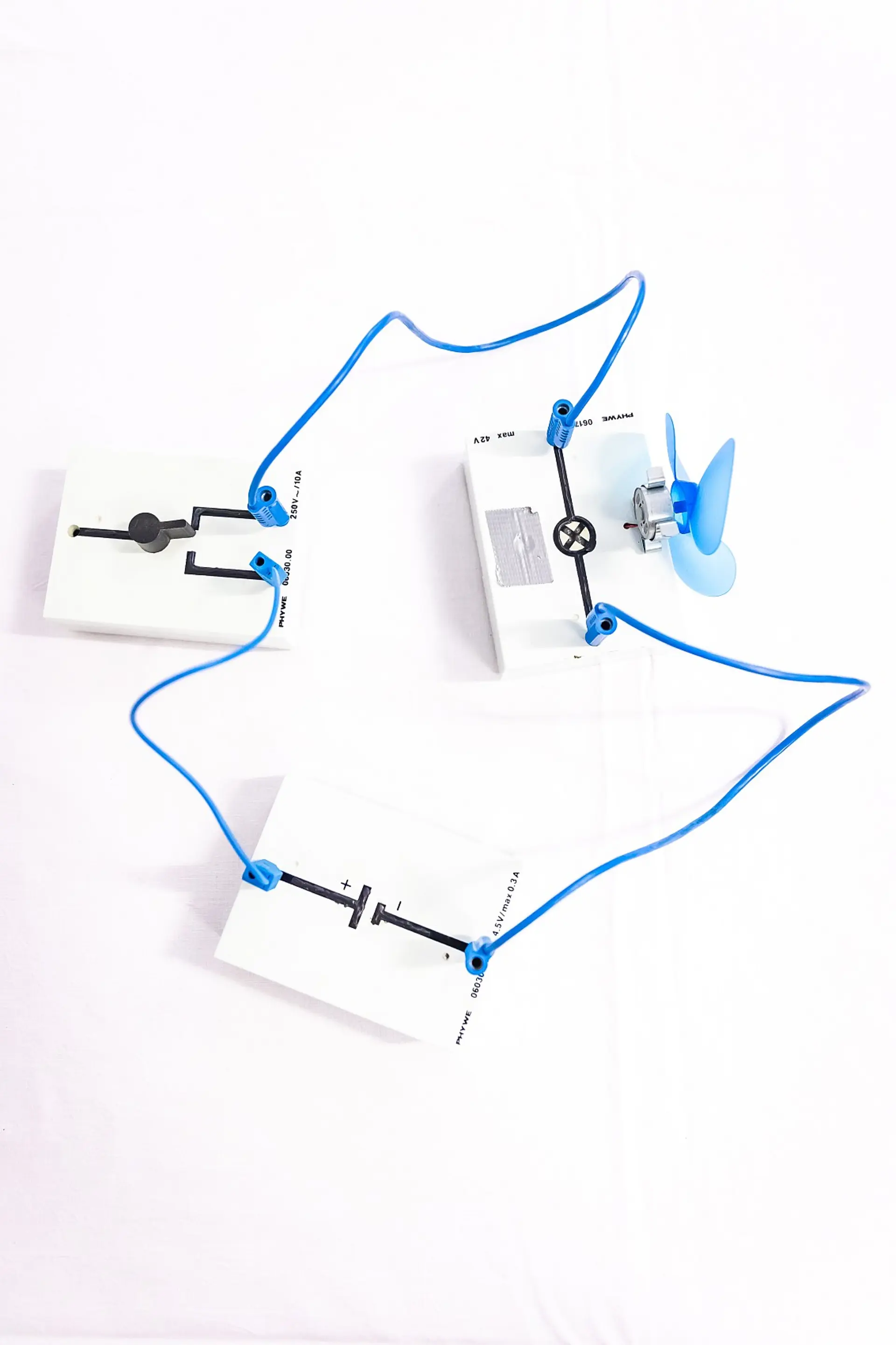 Ein einfacher Stromkreisaufbau auf einer weißen Fläche. Zu sehen sind vier weiße Kunststoffplatten mit aufgedruckten schwarzen Stromsymbolschaubildern. Sie sind über blaue Kabel miteinander verbunden.