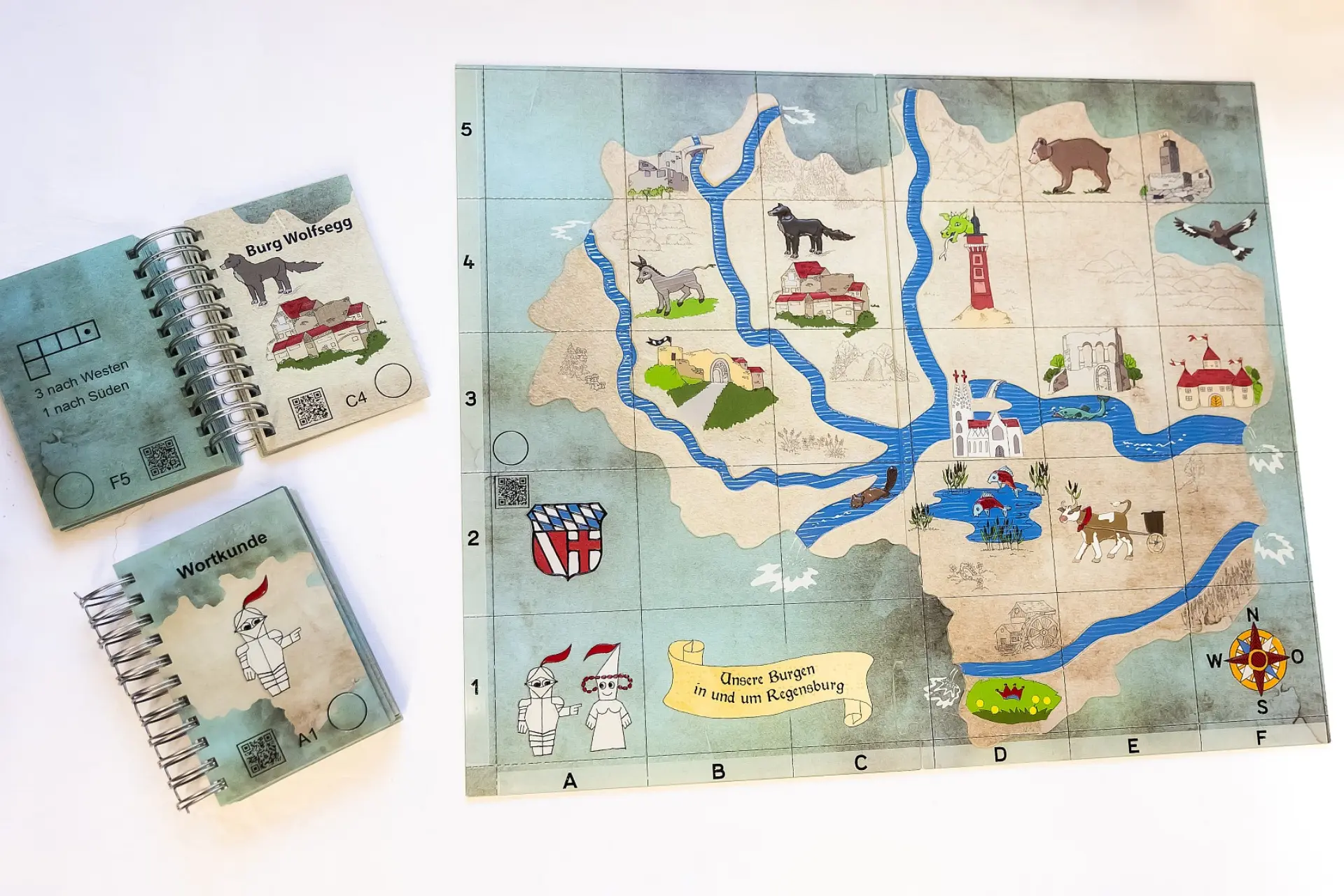 Ein Lehrmaterial-Set für sehbehinderte Kinder, bestehend aus einer großen, taktilen Landkarte von Burgen in und um Regensburg sowie zwei kleineren, spiralgebundenen Büchern. Die Karte zeigt Flüsse, Burgen, Tiere und Gitterlinien mit Koordinaten (A-F, 1-5). Eines der Bücher hat den Titel 'Burg Wolfsegg' und enthält eine Abbildung der Burg sowie Navigationsanweisungen ('3 nach Westen', '1 nach Süden') und QR-Codes. Das andere Buch ist mit Wortkunde beschriftet und zeigt eine stilisierte Figur. Alle Materialien sind taktil und farbenfroh gestaltet.