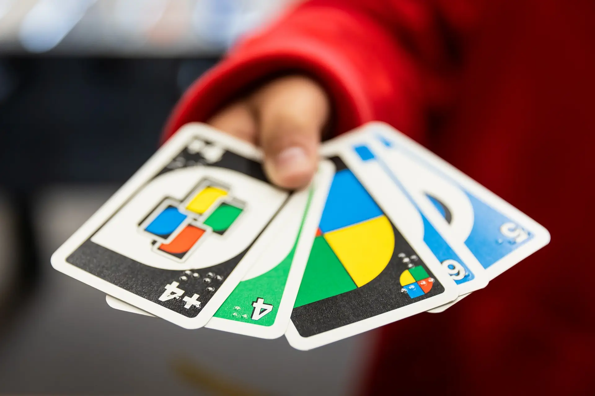 Eine Kinderhand hält mehrere Uno-Spielkarten. Auf jeder Karte sind sowohl die klassischen Uno-Zahlen und -Symbole in Rot, Grün, Blau oder Gelb, als auch die Zahlen in Braille-Schrift geschrieben.