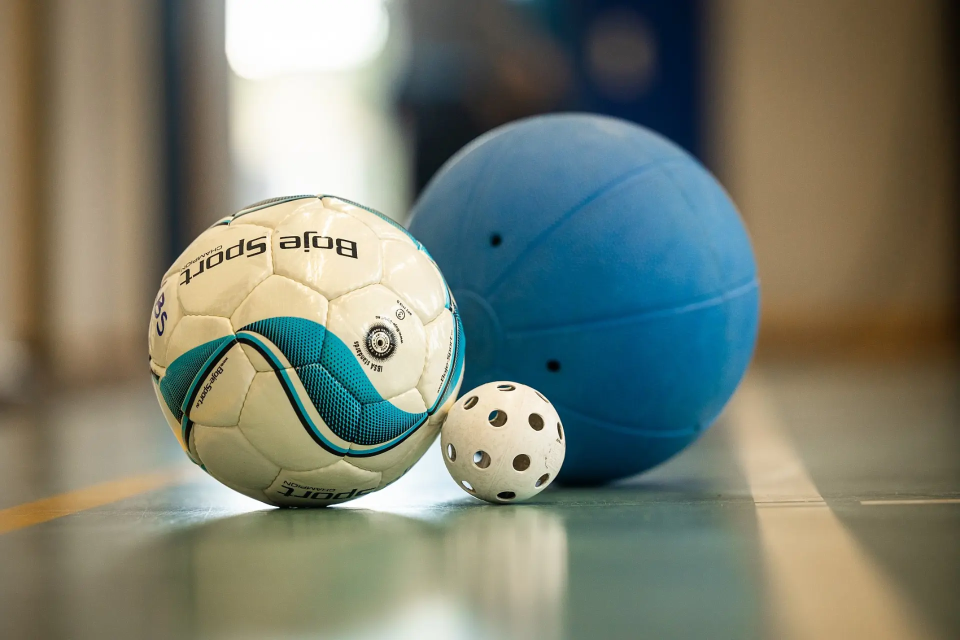 Drei verschiedene Sportbälle für Menschen mit Blindheit oder Sehbehinderung liegen auf einem Hallenboden: Ein weiß-blauer Fußball mit der Aufschrift „Bore Sport“, ein kleiner, weißer Ball mit Löchern (für Torball oder Blindenfloorball) und ein großer, blauer Ball für Goalball.