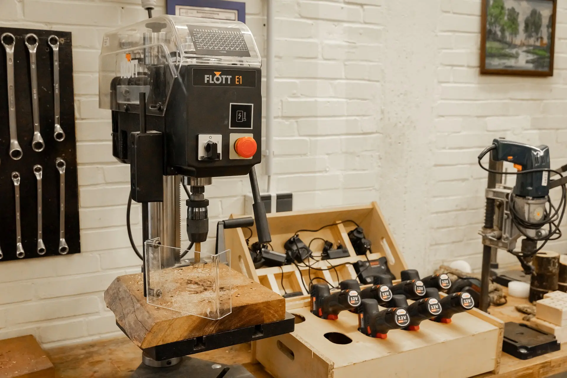 Eine Werkbank in unserer Werkstatt. Im Vordergrund steht eine Standbohrmaschine der Marke "Flott E1" mit einem Stück Holz eingespannt. Die Maschine hat eine Schutzhaube und gut sichtbare Bedienelemente, darunter einen großen roten Not-Aus-Knopf. Im Hintergrund sind weitere Werkzeuge zu sehen, die Umgebung ist ordentlich..