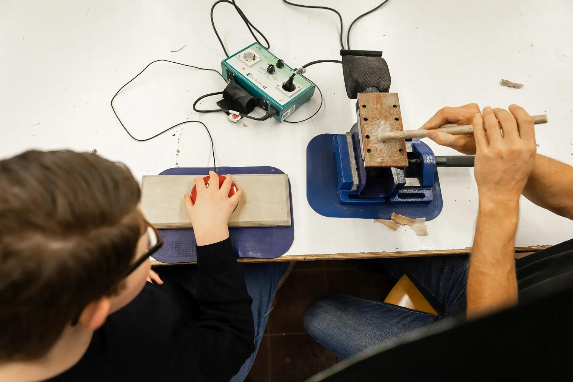 Zwei Personen arbeiten an einem Werktisch: Links sitzt ein Schüler mit Brille und setzt seine rechte Hand auf einen handgroßen Bedienknopf, während eine zweite Person (ein Erwachsener) rechts ein rundes Werkstück in einen blauen Schraubstock eingespannt hält und daran mit einem Werkzeug arbeitet. Auf dem Tisch steht ein grünes Steuergerät mit Kabeln, das vom Schüler bedient wird.