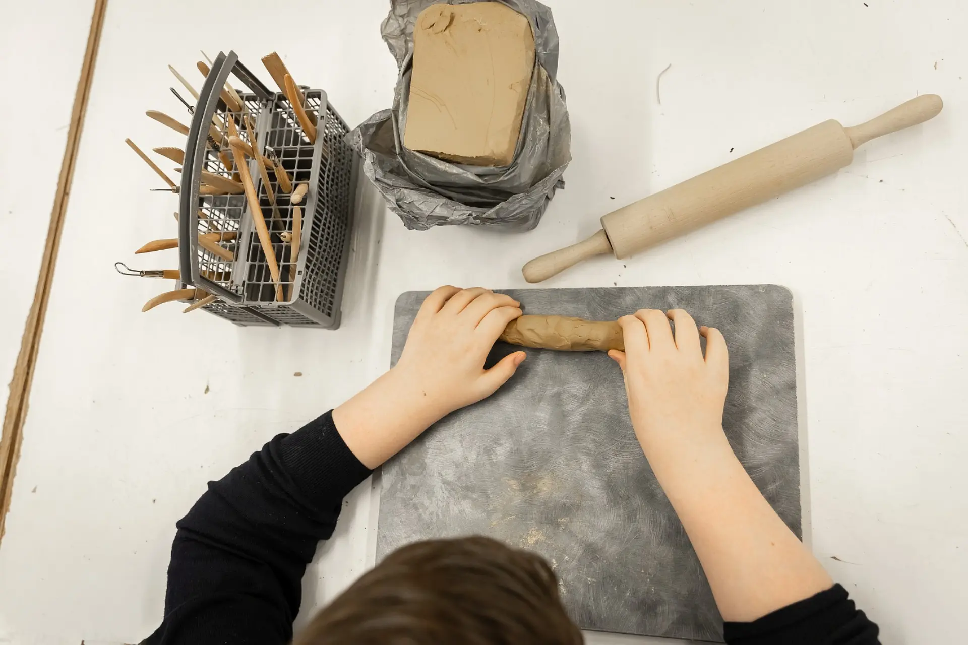Die Hände eines Schülers, der einen Stück rohe Tonmaße auf einem Brett knetet. Neben dem Brett liegen noch ein Block unverarbeitetes Tons, ein Nudelholz sowie ein Behälter mit Werkzeugen zum Schneiden und zu Gravur des Tons.