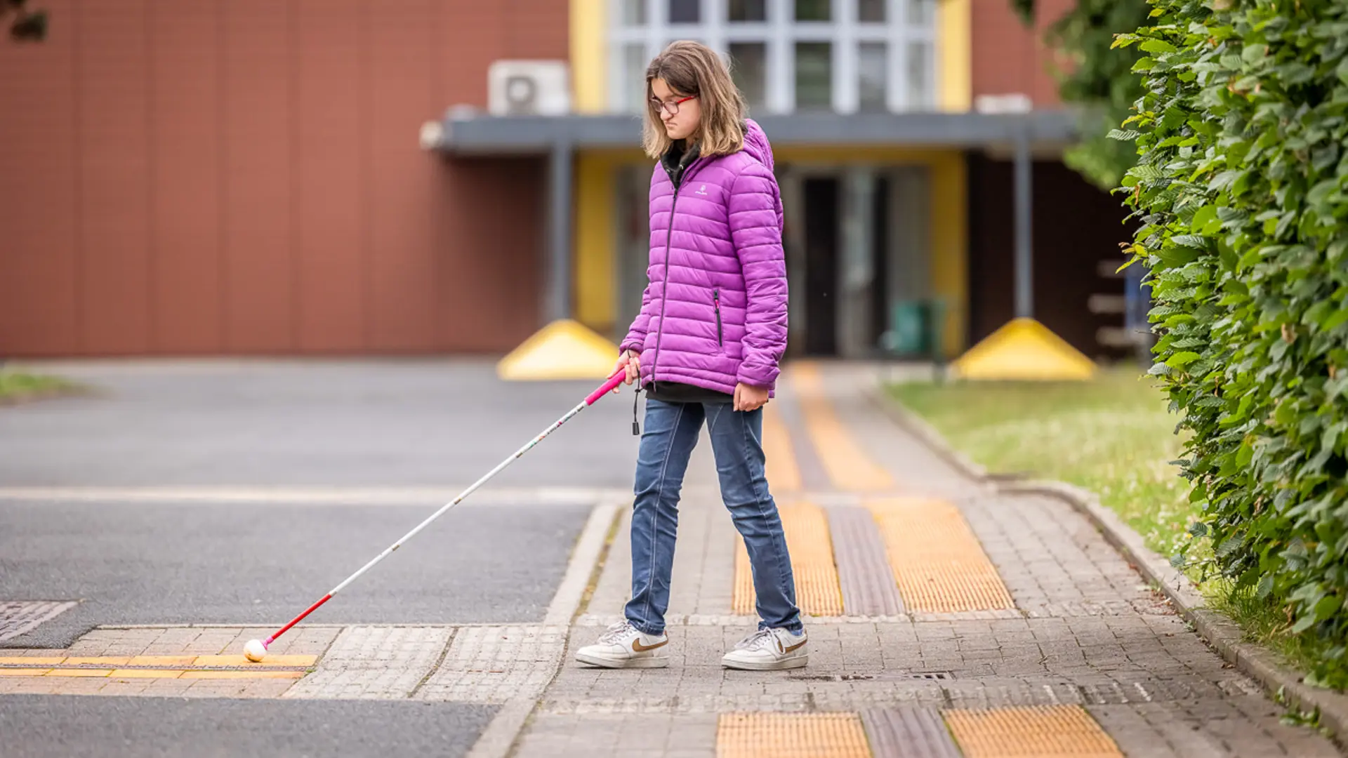 Eine Schülerin mit Blindheit läuft auf dem Weg zwischen den Schulgebäuden und orientiert sich mit einem Blindenstock anhand der Markierungen auf dem Boden. 