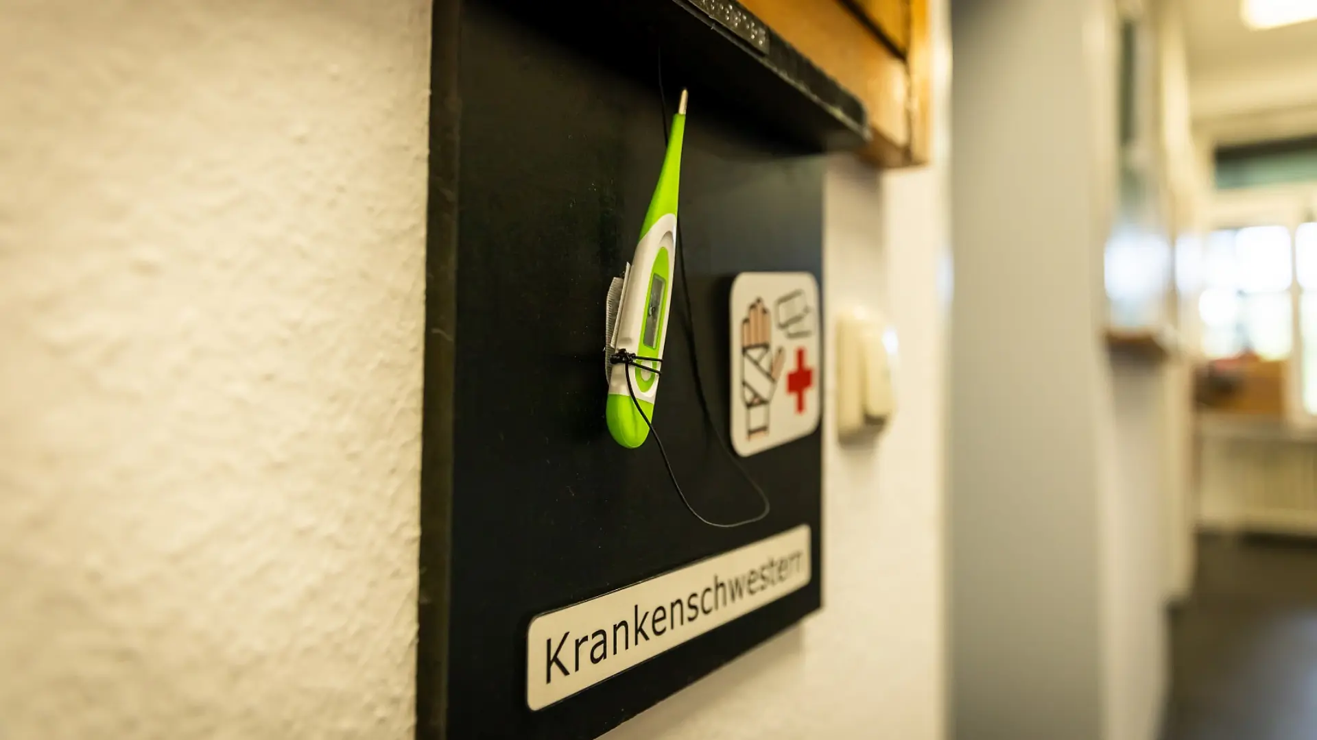 Eine Tafel an der Wand neben einem Zimmer im Schulgebäude. Darauf steht der Text "Krankenschwester". Dazu sind darauf noch ein Fiebermesser aus Plastik sowie das passende Metacom-Symbol zu ertasten bzw. zu sehen. 
