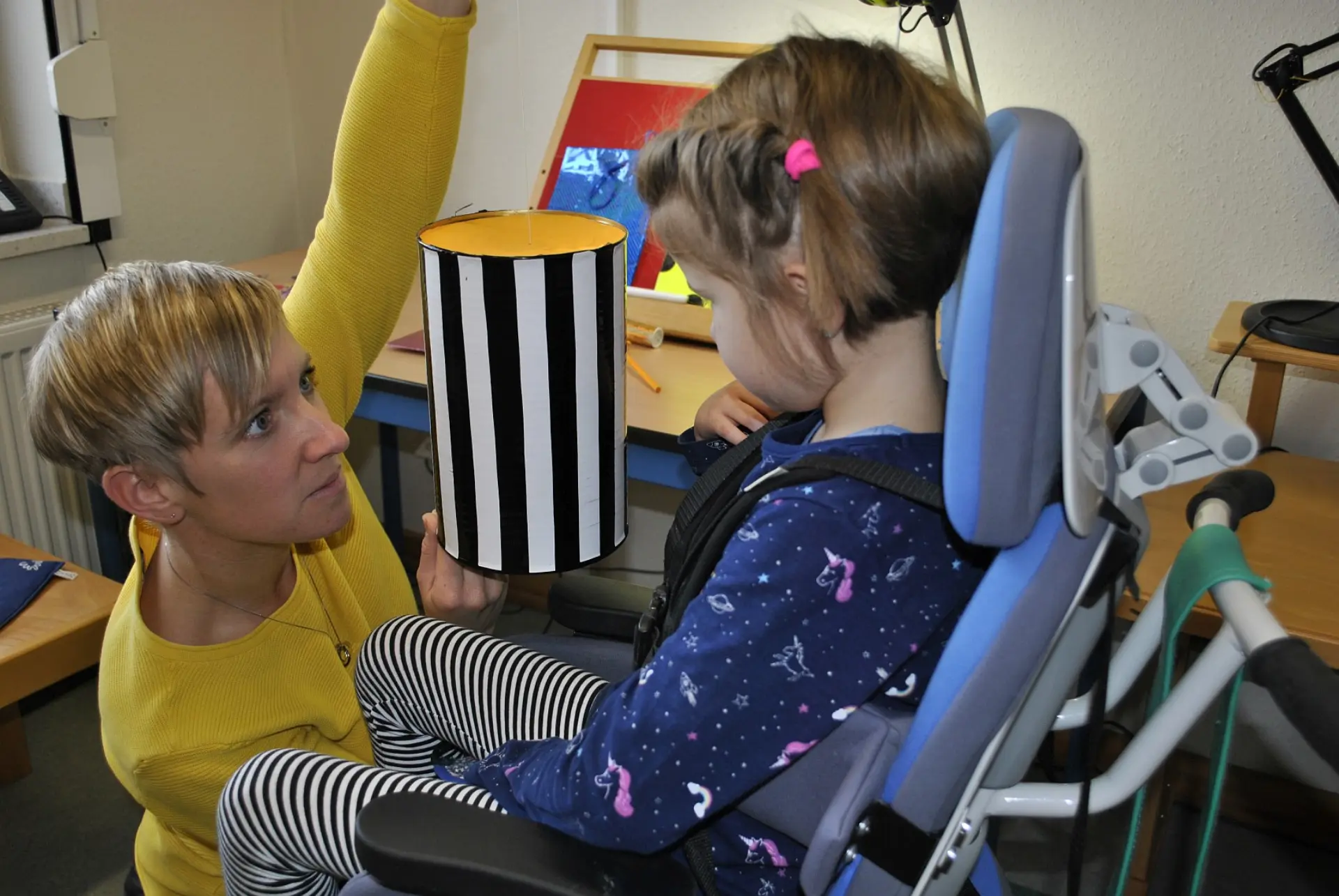 Eine Fachperson steht von einem Mädchen und zeigt ihm eine schwarzweiß-gestreifte Box. Die Fachkraft beobachtet dabei sehr konzentriert das Kind, da sie so eine Sehvermögen-Überprüfung durchführt.