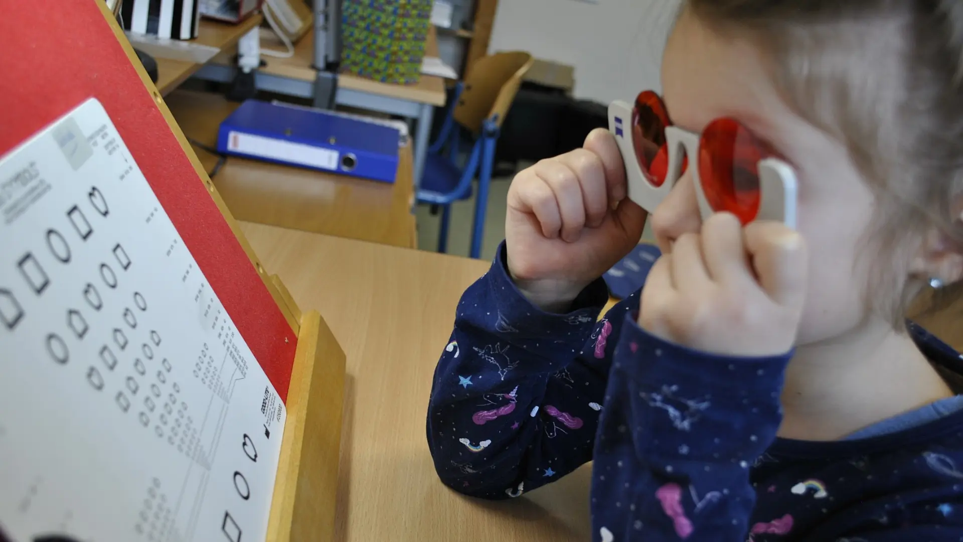 Ein Mädchen hält eine Polarisationsbrille vor ihren Augen und schaut dabei zu einem Augentestblatt mit verschiedenen Formen in unterschiedlichen Größen. Die Formen sind in Reihen und nur als schwarze Linien zu sehen.