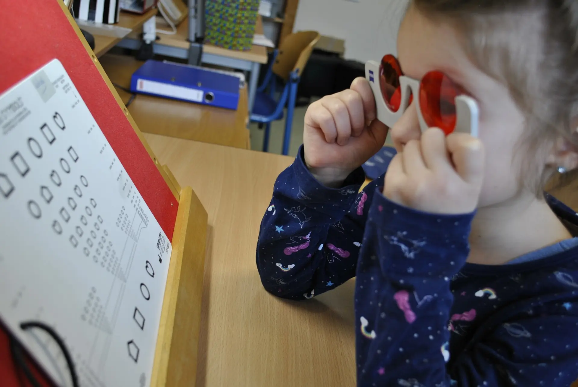Ein Mädchen hält eine Polarisationsbrille vor ihren Augen und schaut dabei zu einem Augentestblatt mit verschiedenen Formen in unterschiedlichen Größen. Die Formen sind in Reihen und nur als schwarze Linien zu sehen.