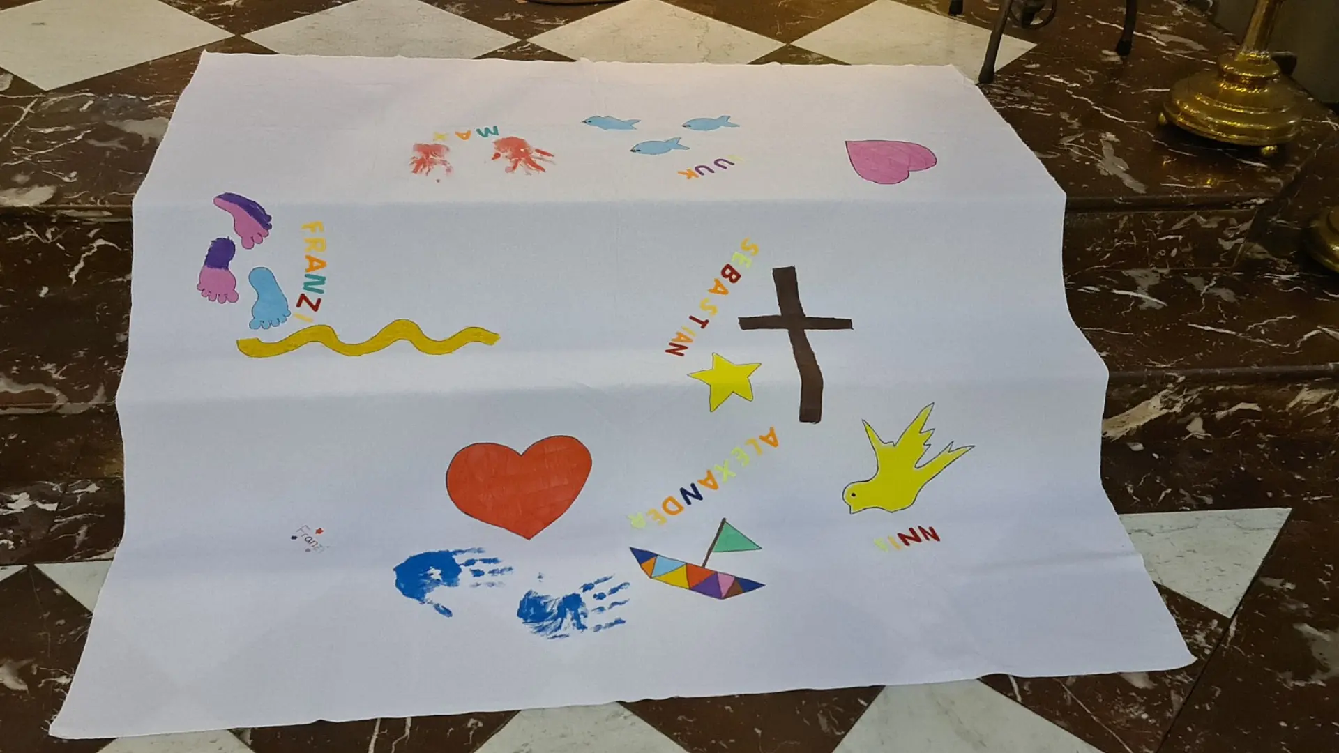 Drei Marmortreppen in einer Kirche, die zum Altar führen. Darauf liegt ein großer weißer Stoff, auf dem die Namen der getauften Kinder sowie christliche Elemente wie ein Kreuz, ein Herz, eine Taube gemalt sind.