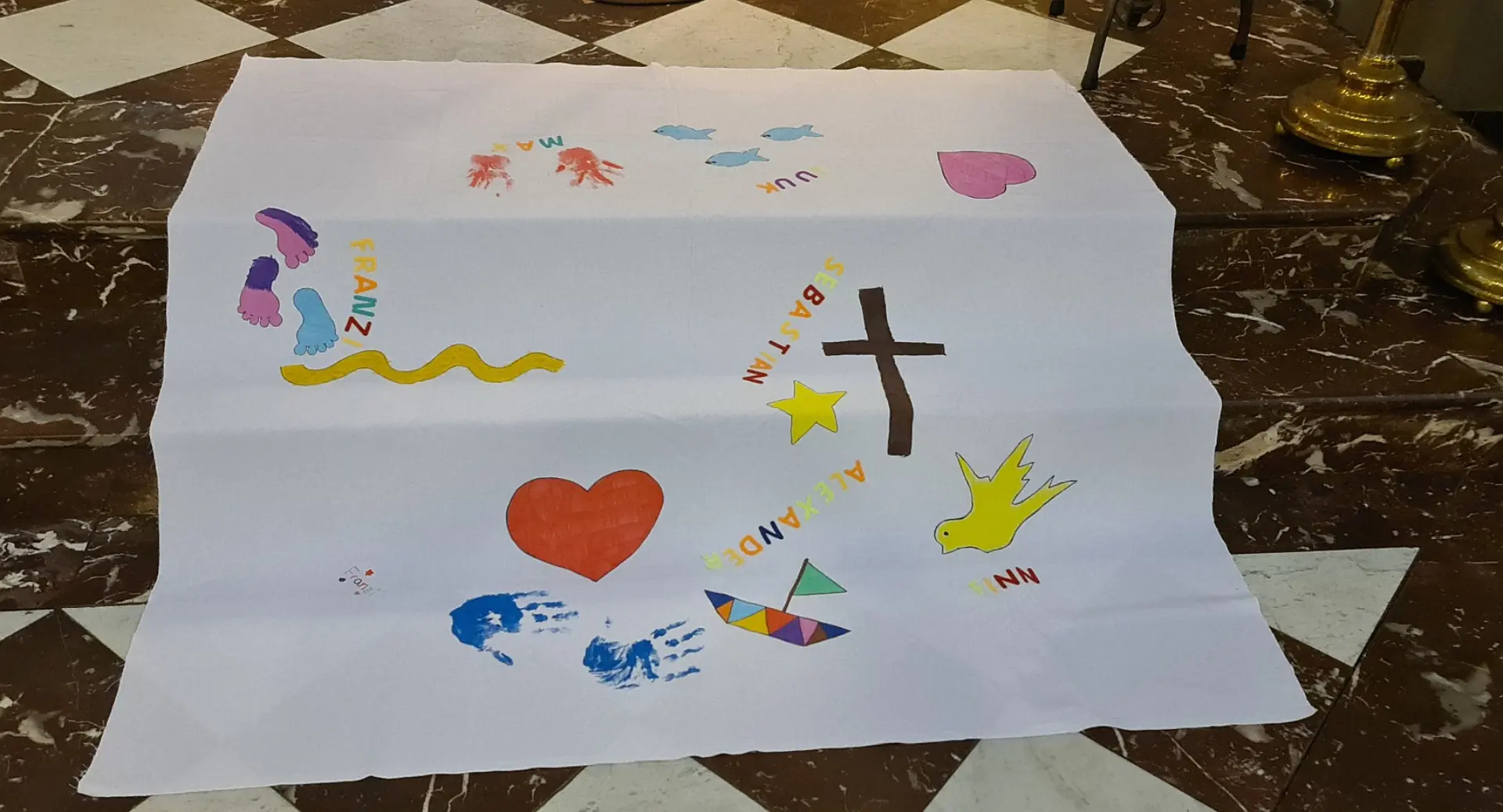 Drei Marmortreppen in einer Kirche, die zum Altar führen. Darauf liegt ein großer weißer Stoff, auf dem die Namen der getauften Kinder sowie christliche Elemente wie ein Kreuz, ein Herz, eine Taube gemalt sind.