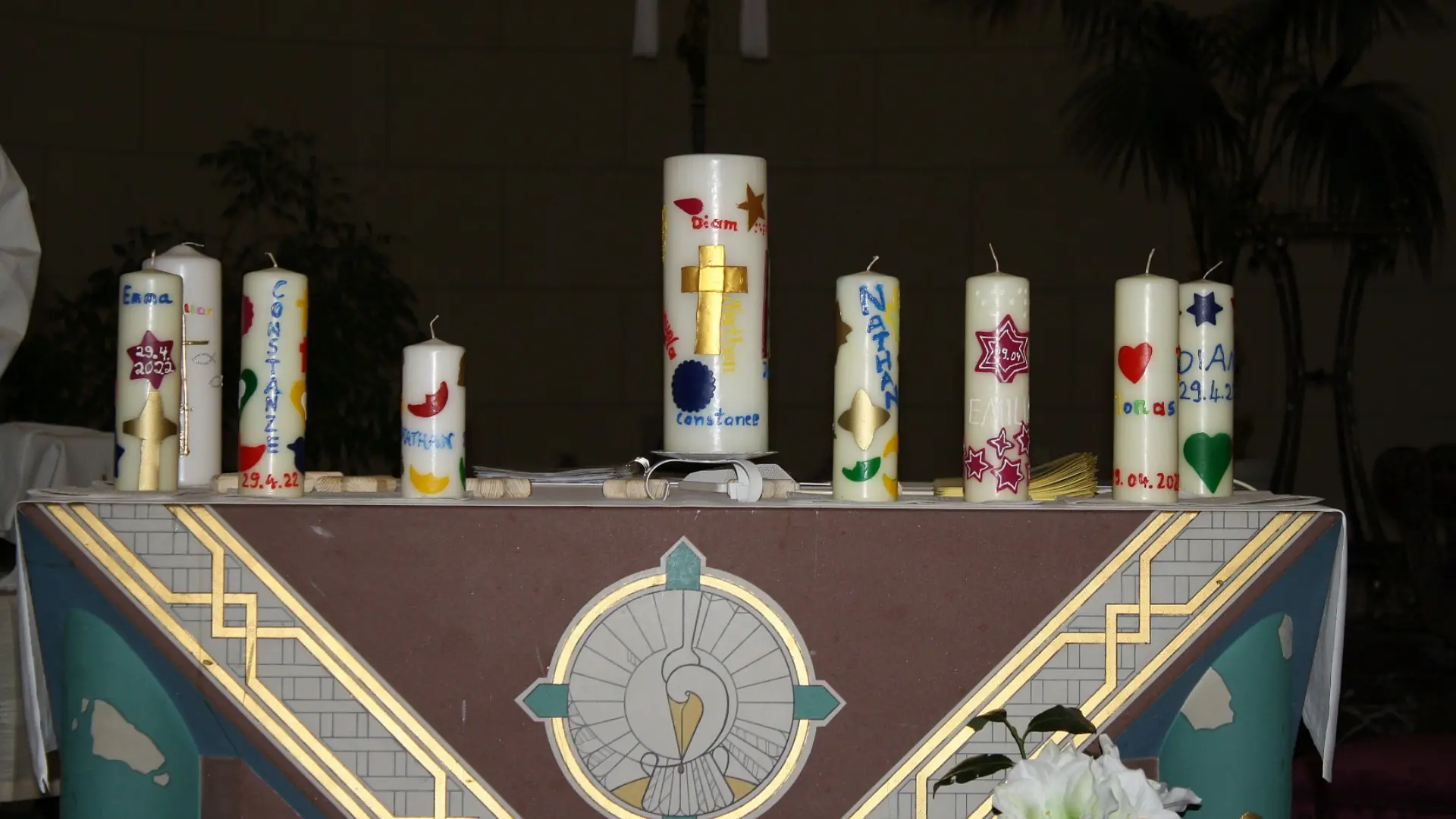 Ein Tisch mit einer festlichen Tischdecke mit goldenen christlichen Ornamenten verziert. Auf dem Tisch sind acht mittelgroße und eine größere Taufkerzen zu sehen, alle sind mit bunten Elementen wie Sternen, Monden und Herzen sowie mit den Namen der getauften Kindern bemalt.