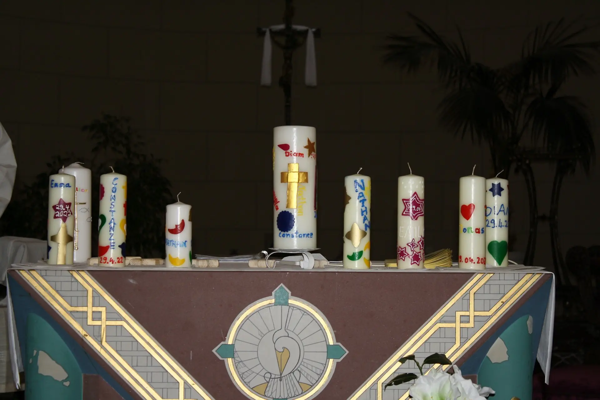 Ein Tisch mit einer festlichen Tischdecke mit goldenen christlichen Ornamenten verziert. Auf dem Tisch sind acht mittelgroße und eine größere Taufkerzen zu sehen, alle sind mit bunten Elementen wie Sternen, Monden und Herzen sowie mit den Namen der getauften Kindern bemalt.