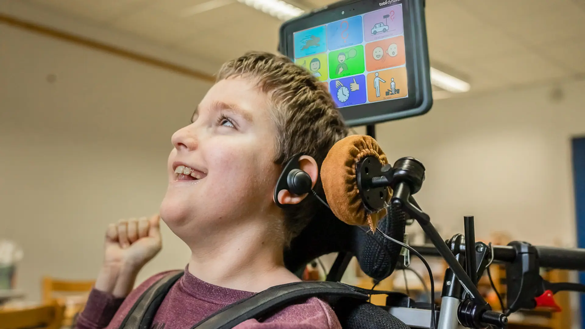 Ein Schüler mit Komplexer Beeinträchtigung sitzt im Rollstuhl und lacht. An seinem Ohr und neben seiner linken Gesichtsseite ist ein Kopftaster angebracht, über seinem Kopf ist ein kleiner Monitor mit Metacom-Symbolen. Der Schüler kommuniziert mittels auditiven Scannings. 