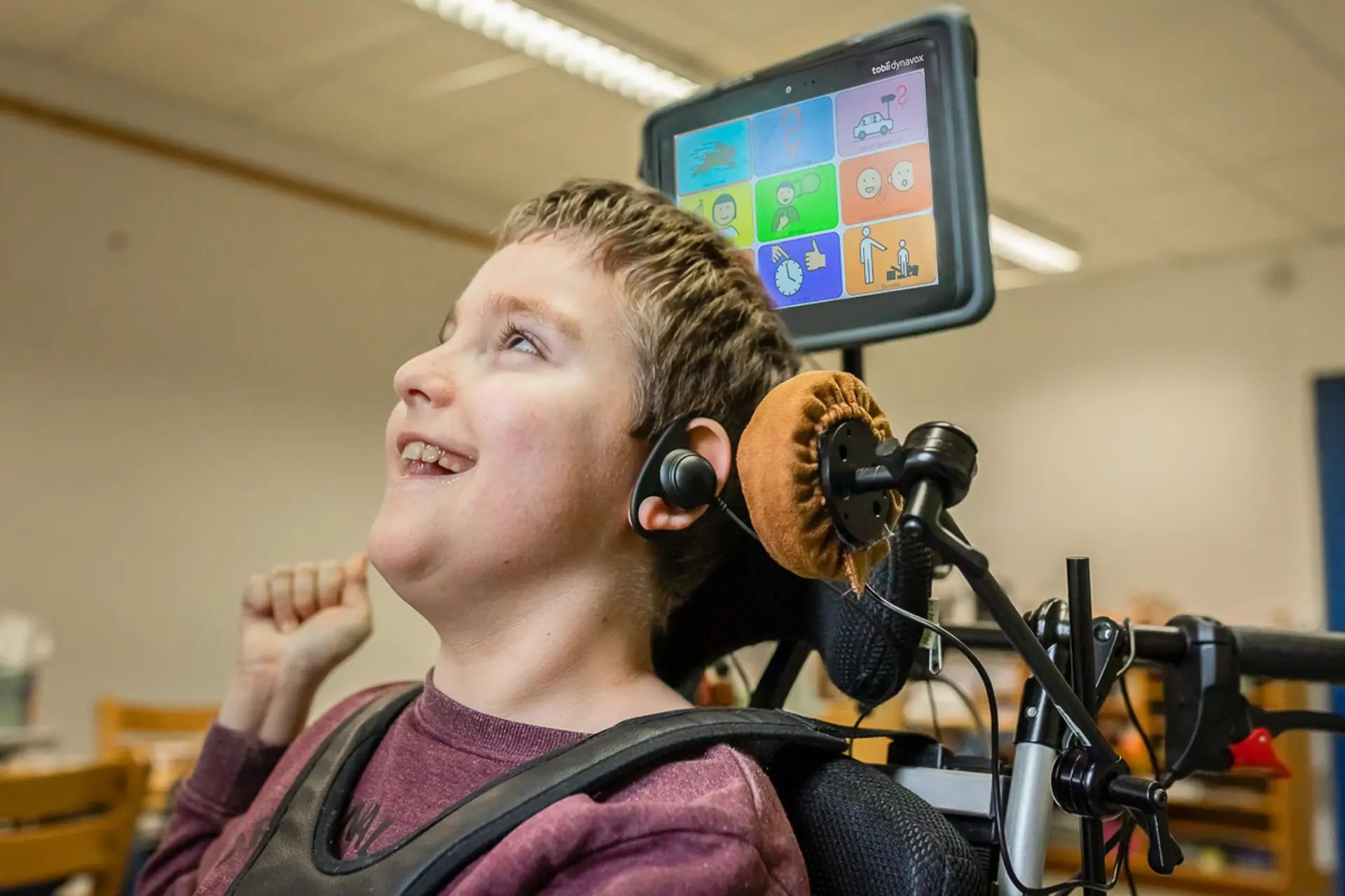 Ein Schüler mit Komplexer Beeinträchtigung sitzt im Rollstuhl und lacht. An seinem Ohr und neben seiner linken Gesichtsseite ist ein Kopftaster angebracht, über seinem Kopf ist ein kleiner Monitor mit Metacom-Symbolen. Der Schüler kommuniziert mittels auditiven Scannings. 