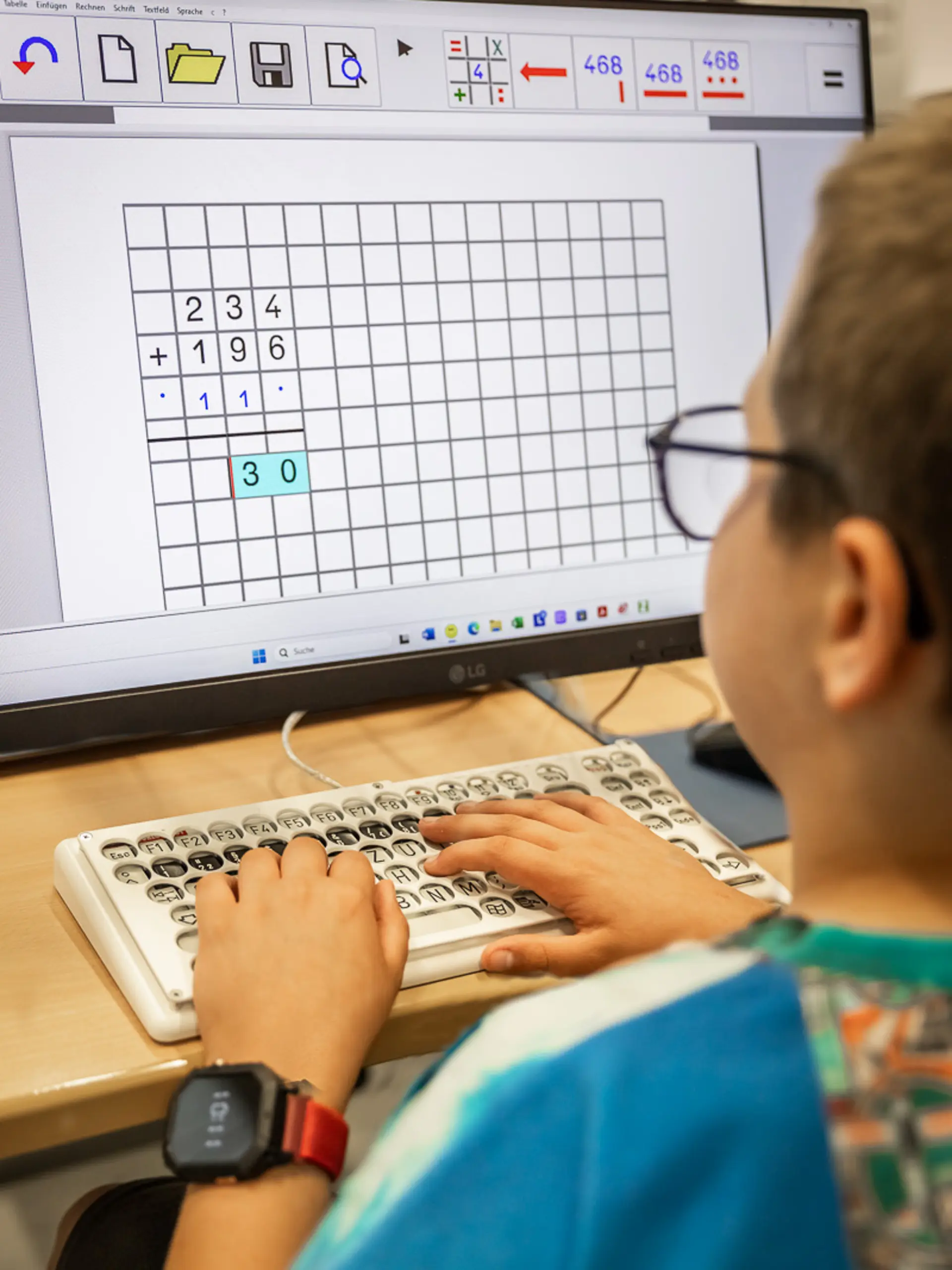 Ein Schüler sitzt am PC in einem Klassenzimmer und blickt zum Computermonitor. Er benutzt eine Spezialtastatur und macht Matheaufgaben.