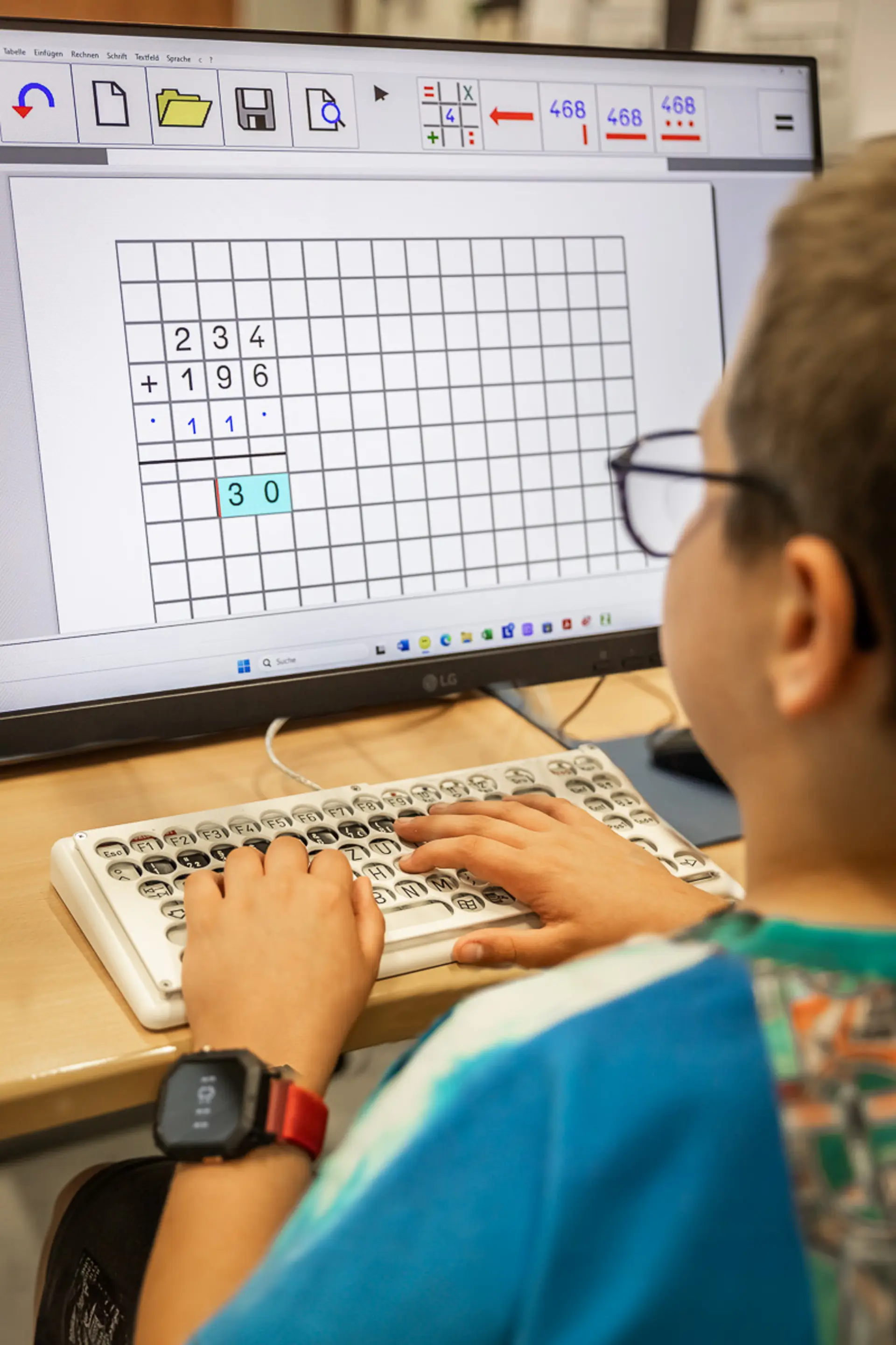 Ein Schüler sitzt am PC in einem Klassenzimmer und blickt zum Computermonitor. Er benutzt eine Spezialtastatur und macht Matheaufgaben.